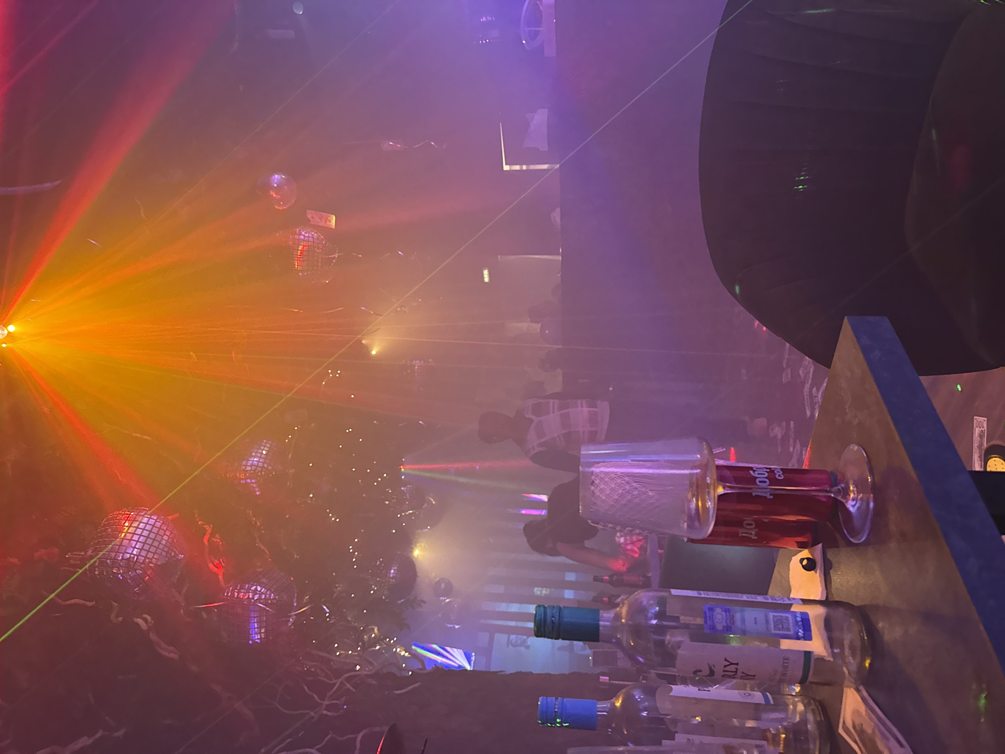 The image shows the interior of a bar or nightclub in what is likely Bobrovo, Russia. The scene is illuminated by colorful lighting, including orange, purple, and green laser beams. Disco balls are suspended from the ceiling, reflecting light throughout the space.
In the foreground, a bar top is visible. Empty wine bottles and glasses are scattered across the surface. Some bills are also visible.
In the background, several people stand near the bar. One individual wears a checkered shirt. The scene has a party or nightlife atmosphere.