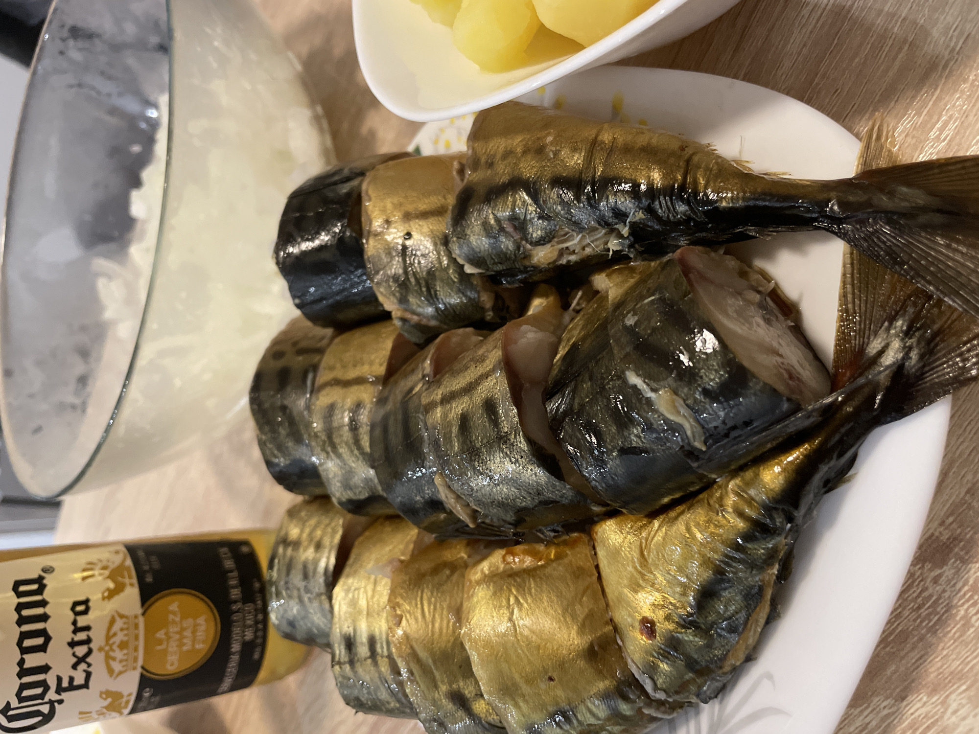 An overhead, medium shot captures a domestic scene, likely in Moscow, Russia, featuring a plate of sliced smoked mackerel, a bottle of Corona Extra beer, a bowl of chopped onions, and a bowl of boiled potatoes. The mackerel, appearing to be whole fish cut into sections, are arranged on a white plate with a subtle yellow floral pattern. Some pieces display a golden, oily sheen, while others have darker, more pronounced skin. The Corona Extra bottle, with its distinctive yellow label, stands to the left of the plate. Behind the fish, a large glass bowl contains a generous amount of finely chopped white onions. To the right of the plate, a white bowl holds cubed, bright yellow boiled potatoes. The scene is set on a light-colored wooden table. No individuals are visible in the frame.