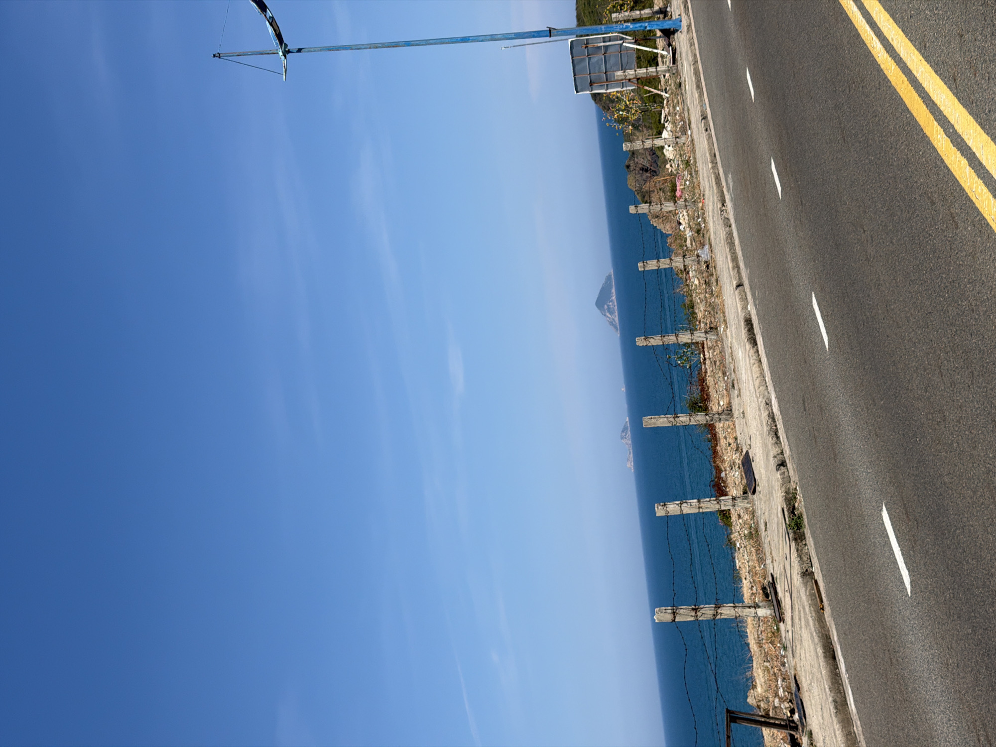 A rotated eye-level shot reveals a road with double yellow lines running along its right edge, parallel to the sea. A rustic barrier of concrete posts and barbed wire separates the road from the sea. A signpost is visible near the barrier. A distant, pyramid-shaped island rises from the water. The sky is blue with some clouds. This scene is located along the Bac Nha Trang Ward road, Vietnam.