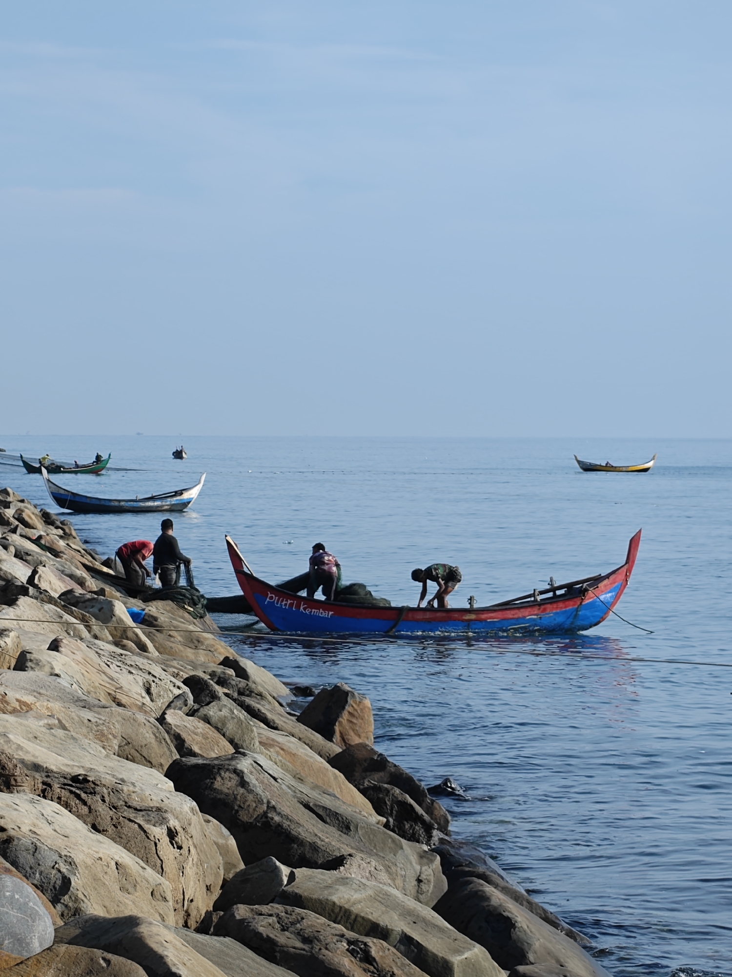 The photograph captures a tranquil coastal scene in Lhokseumawe, Indonesia. Several traditional fishing boats, brightly colored, are visible in the calm, light blue waters. Three men are working on one boat, which is secured near a rocky embankment. The boat is painted blue and red with the words "PUTRI KEMBAR" on its side. Two other men are positioned near a boat along the rocky embankment, attending to nets. In the distance, more boats are scattered across the water, and the sky is a clear, pale blue. The foreground is dominated by large, gray rocks that form the coastline, leading to a serene and somewhat remote maritime setting.