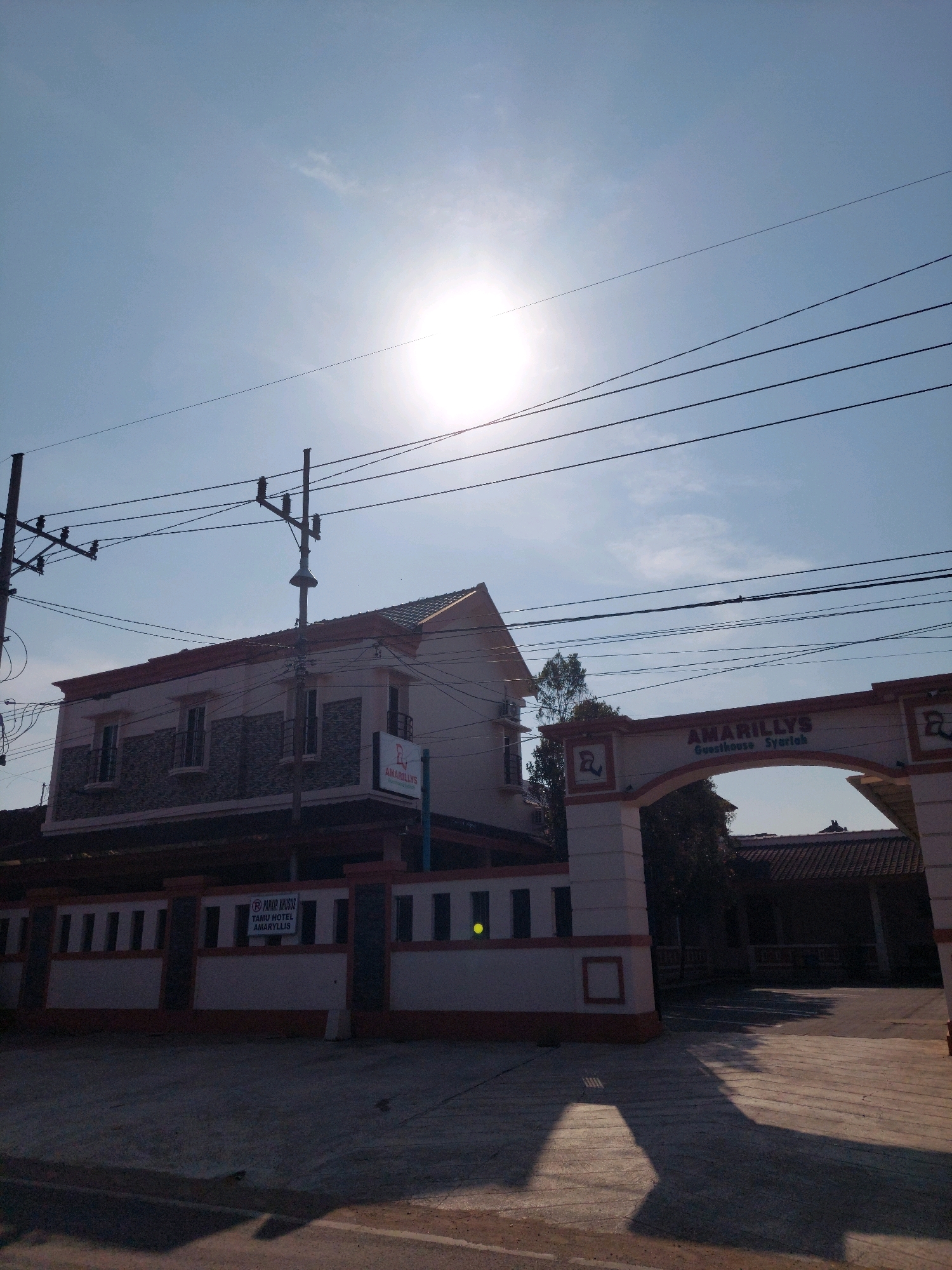 The image depicts the entrance to Amaryllis Guesthouse Syariah in Menadi, Indonesia. The sun is high in the sky, casting long shadows across the paved ground. Overhead, a network of power lines crisscrosses the frame. To the left, a two-story building with a tiled roof is visible, featuring stone accents and windows. A sign on the building reads "AMARYLLIS" with a red logo. To the right, an arched gateway marks the entrance, with "AMARYLLIS Guesthouse Syariah" written above the arch. Adjacent to the gateway is a smaller building with a dark tiled roof. A sign on a fence to the left of the building states "PARKIR KHUSUS TAMU HOTEL AMARYLLIS." The overall scene suggests a bright, sunny day at the guesthouse.