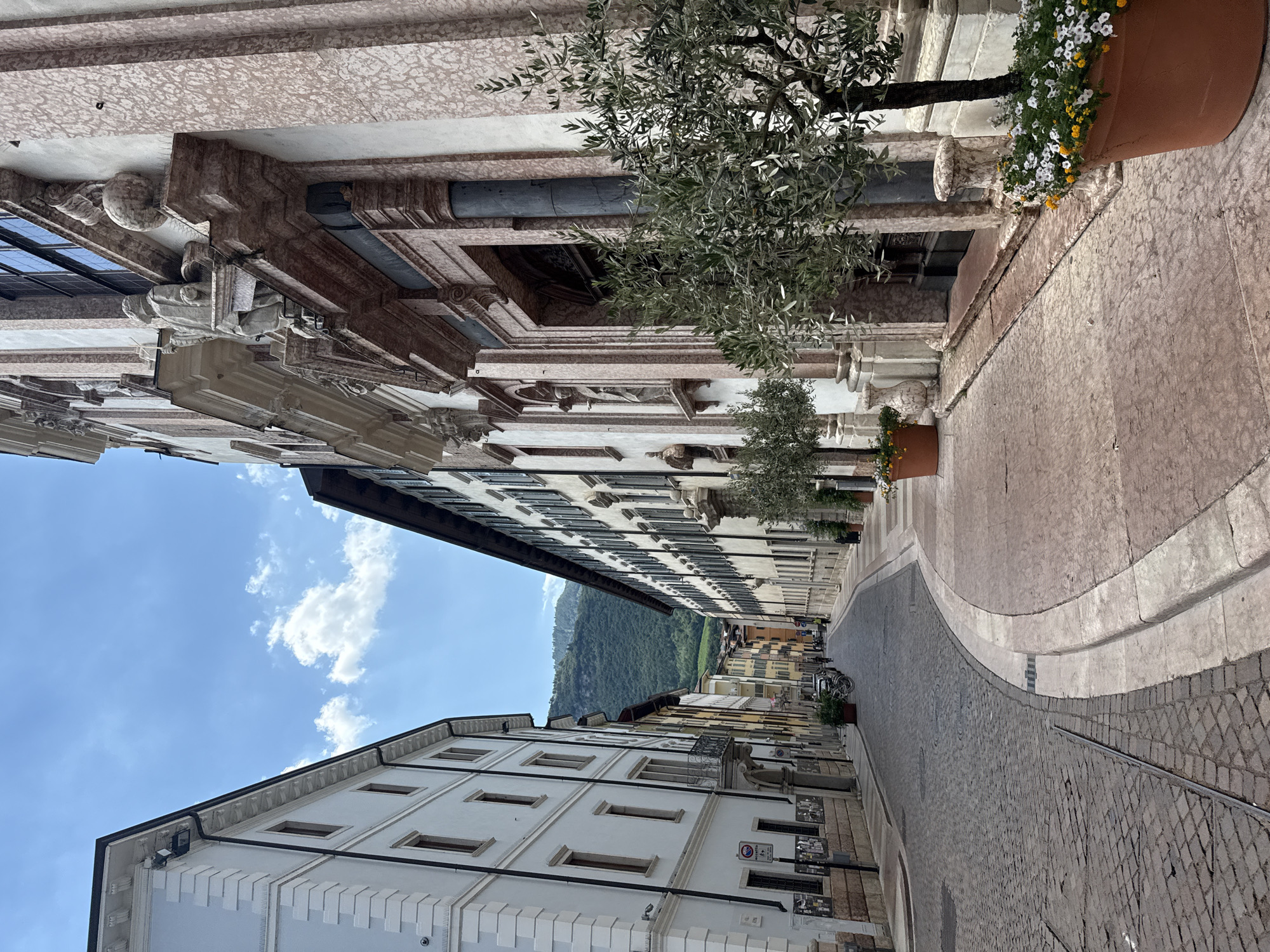 The image depicts a street scene in Trento, Italy. On the right, a large, ornate building with marble facade and classical architectural details is visible. In front of this building, two potted olive trees with small white and yellow flowers are placed. The street itself is paved with cobblestones. To the left, a narrow street lined with tall, pale buildings with windows and shutters recedes into the distance, with a green, tree-covered mountain visible in the background. The sky is blue with scattered white clouds. Several people are walking in the distance on the left side of the street. A bicycle is parked near the center of the street on the left. A traffic sign and some posters are attached to a wall on the left.