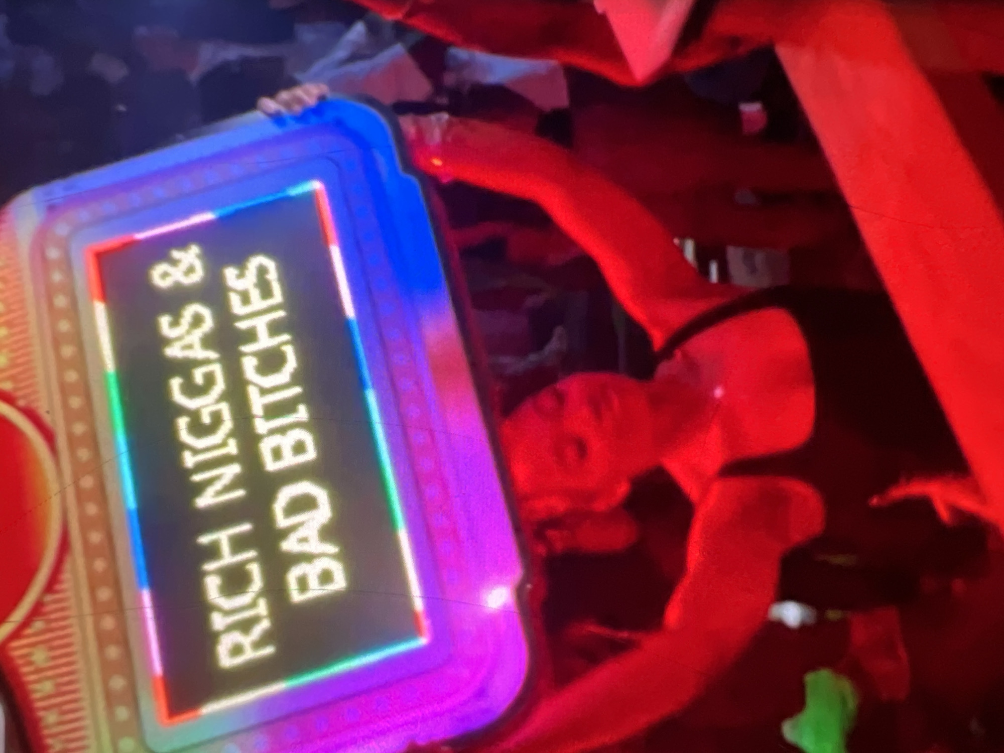 In an indoor setting, possibly a nightclub, a person holds up a large, illuminated sign above their head. The sign displays the words "RICH NIGGAS & BAD BITCHES" in glowing white capital letters, outlined in a rainbow of colored lights. The person holding the sign is partially visible, wearing a black top and appears to be moving their arms. The background is dimly lit, with other people and red lighting suggesting a lively atmosphere. The overall impression is one of a celebratory or provocative event. This scene is not identifiable as being in Lagos, Nigeria, or any other specific city.