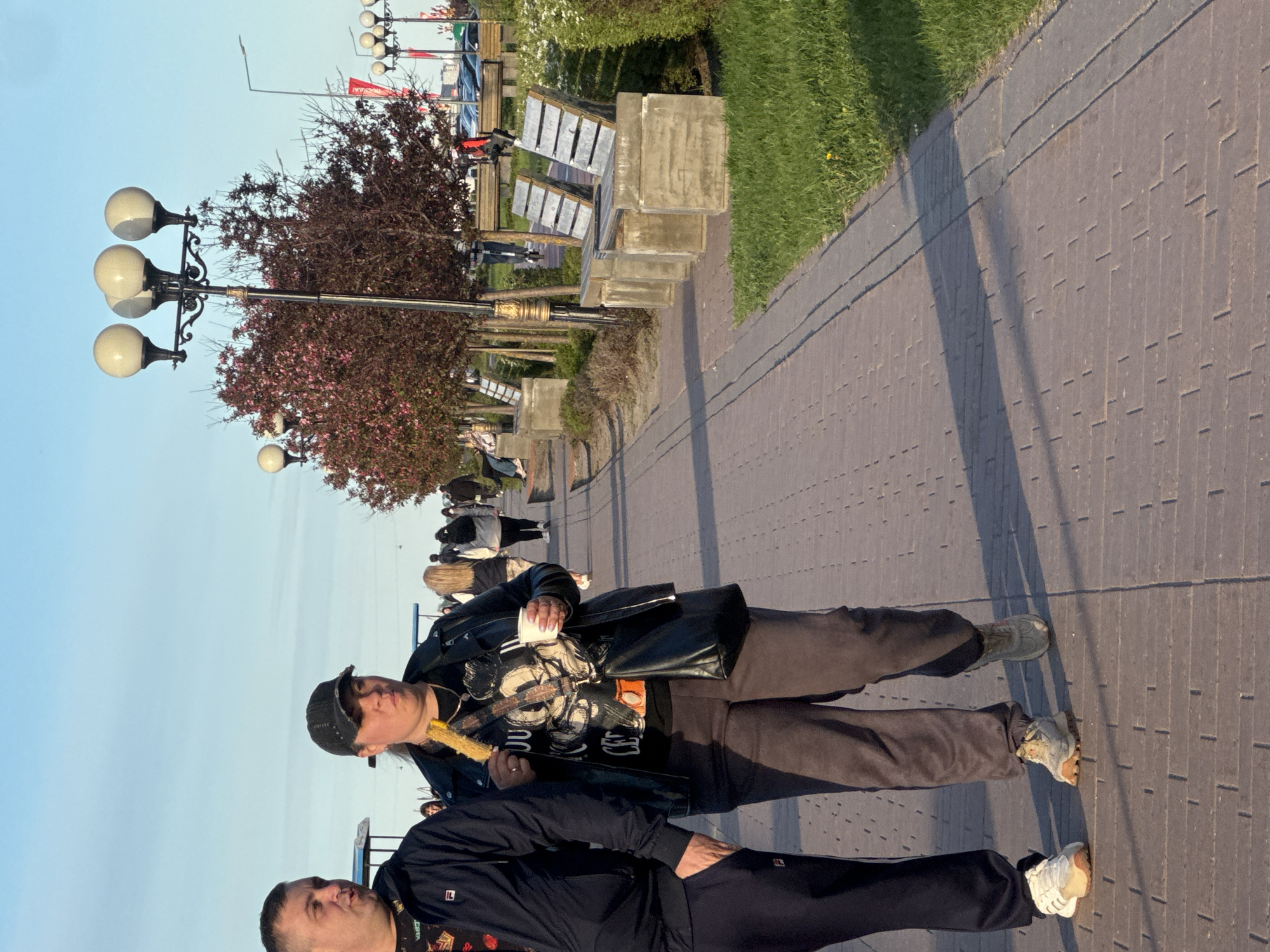 The image shows two people on a brick-paved walkway. The person in the foreground is wearing a black tracksuit. The other person, slightly behind, wears a black leather jacket and grey pants, holding corn on the cob. Both are standing, appearing to converse. In the background, there are benches and street lamps, typical of a public promenade. The presence of the Volga River and benches suggests the Lower Volga Embankment in Nizhny Novgorod, Russia.