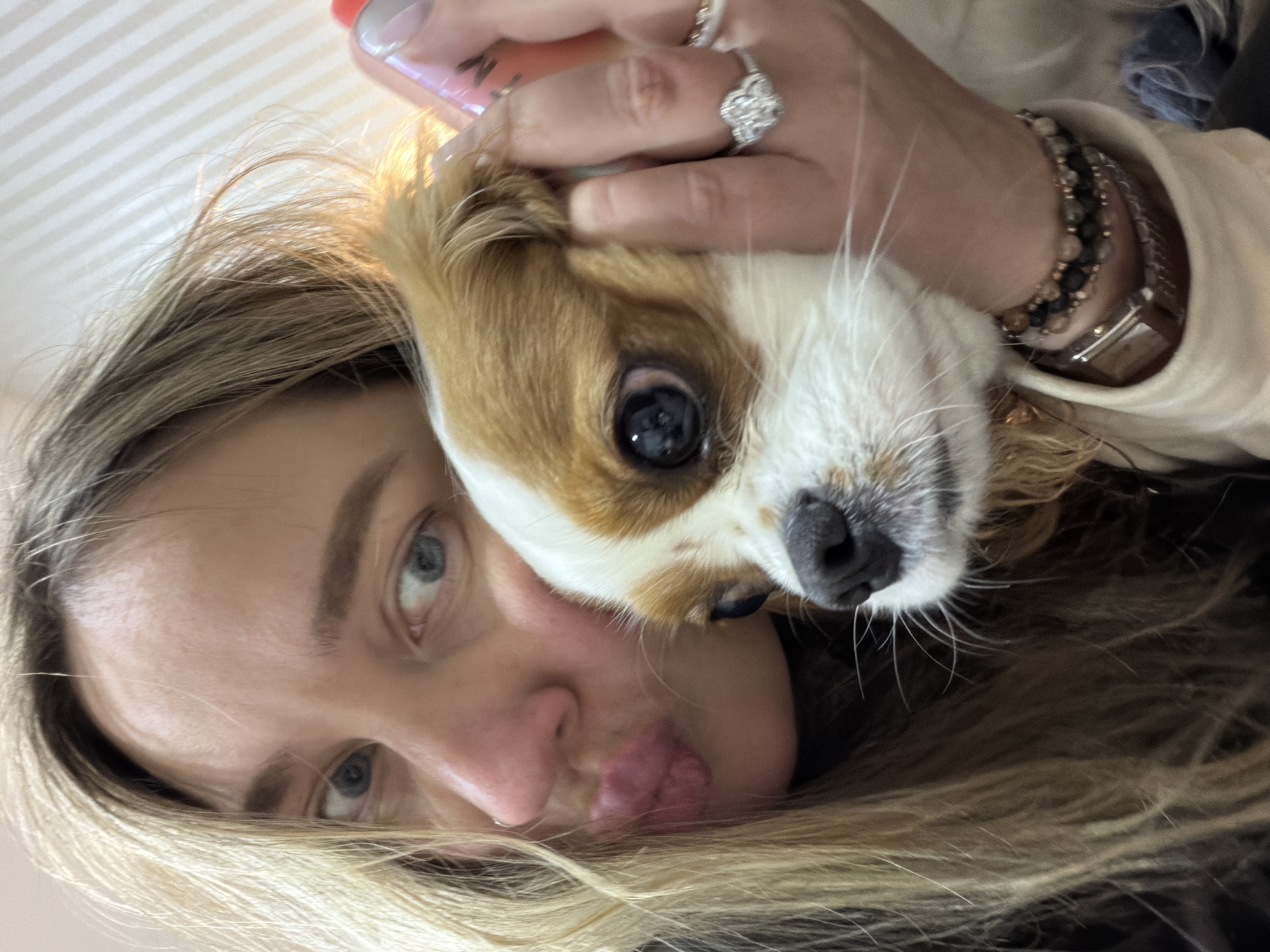 A close-up, low-angle shot shows a person and a small dog in what appears to be an affectionate moment. The image is oriented vertically, with the subjects appearing rotated to the side, looking upwards towards the camera.
The person, seemingly female, has long blonde hair with darker roots, and visible blue eyes. Their face is angled towards the camera, with lips playfully pursed. A small nose piercing is visible on their right nostril. Their hand is seen near the dog's head, adorned with several rings, including a prominent diamond engagement-style ring, and light-colored nail polish with white tips. On their wrist, multiple beaded bracelets and a metallic watch are visible.
The dog is small, with white fur and prominent brown/tan patches, particularly around its eyes and ears. It has dark, round eyes and a black nose with long white whiskers. Its head is resting close to the person's face, suggesting a close bond.
The background consists of a light-colored surface with faint horizontal striped patterns, possibly a ceiling or a textured wall, indicating an indoor setting. A glimpse of a pink object, possibly a phone or bottle, is held by the person's hand.
The overall mood is warm and intimate, focusing on the bond between the person and their pet. No specific time of day or weather can be determined from the image, and there are no discernible location cues within the frame itself to indicate Chaikovsky, Russia. No visible text is present.