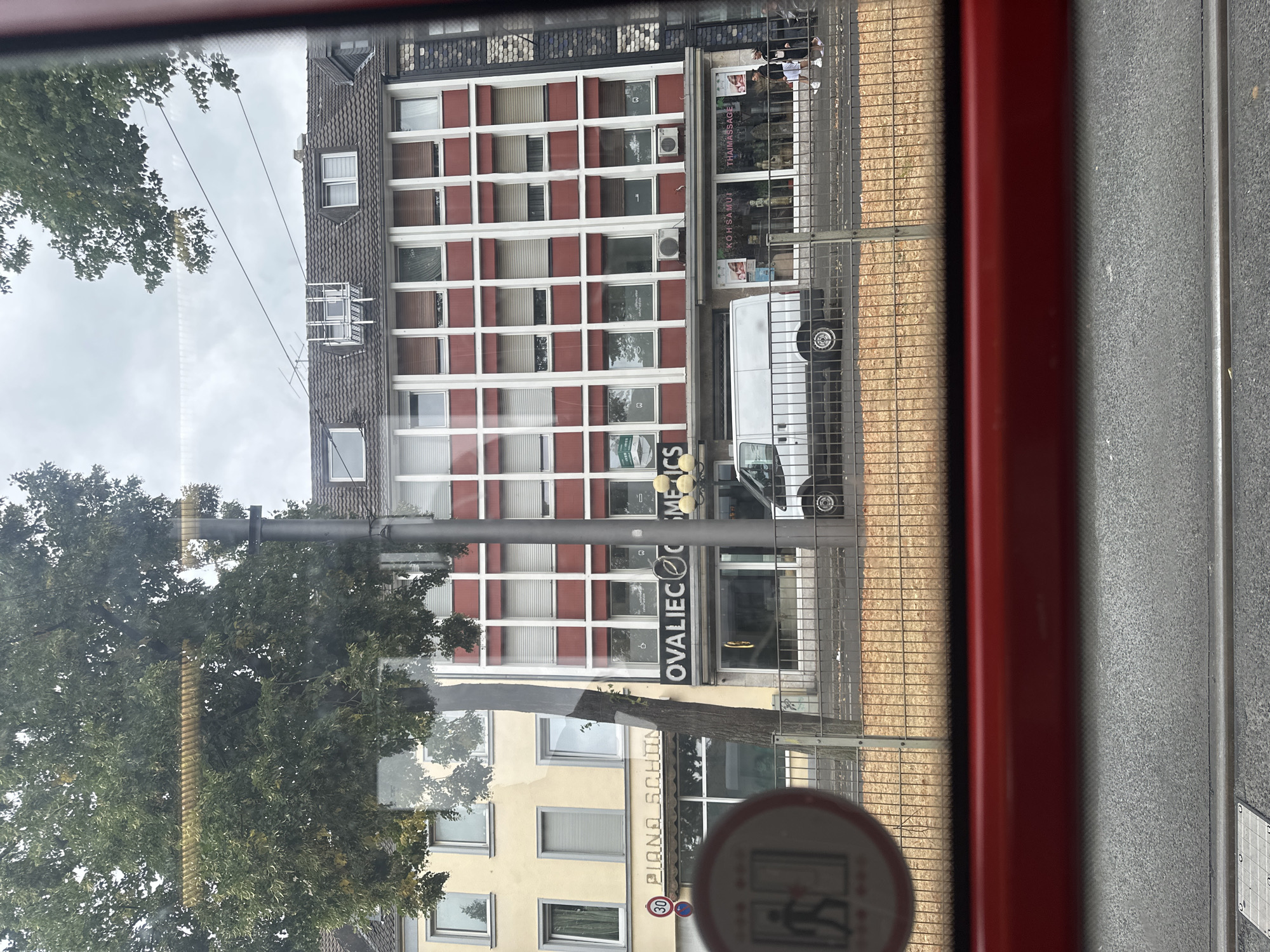 The image shows a street scene in Krefeld, Germany, viewed from inside a vehicle, likely a tram, indicated by the red frame at the bottom and the tracks visible in the foreground. The main focus is on the buildings across the street.
On the left, a light yellow building has multiple windows and a sign reading "PIANO SCHÖN" above a shopfront. A circular sign with a pedestrian icon and arrows is visible on the left side of the street. To the right of this building is a red and white multi-story building with many windows, each covered by blinds. The signage on this building includes "OVALIEC COSMETICS" in large letters, and further to the right, above a shopfront, "KOH SAMUI THAI MASSAGE".
A white van is parked in front of the "KOH SAMUI THAI MASSAGE" establishment. Two women, one in a black jacket and white shorts, and the other in a black jacket and white sneakers, are walking on the sidewalk in front of the massage parlor. A metal fence runs along the sidewalk, separating the street from the buildings. Trees with green foliage are visible on the left side of the image, partially obscuring the buildings. The sky is overcast.