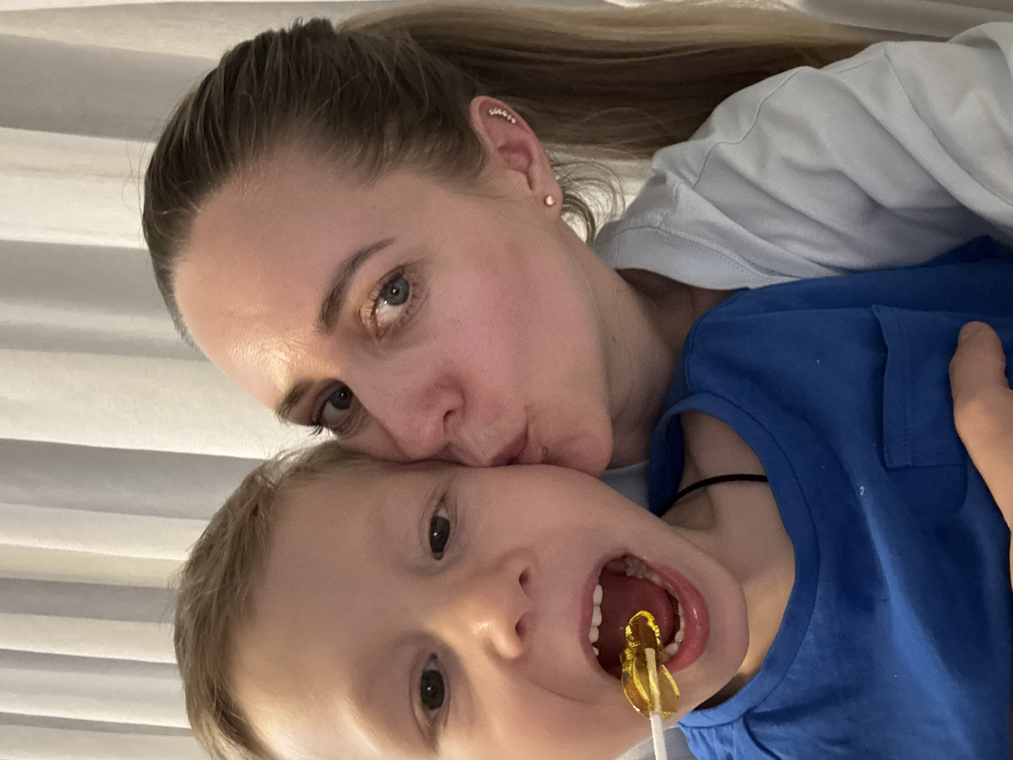 A woman and a child are posed in a selfie. The woman, with her blonde hair pulled back in a ponytail, is kissing the child’s forehead. The child has blond hair and is wearing a blue shirt. The child has an open mouth and a yellow lollipop is inside. The background is blurred and indistinct, appearing to be a room with white horizontal blinds. There is no confirmed location or association with the city of Chaikovsky, Russia.