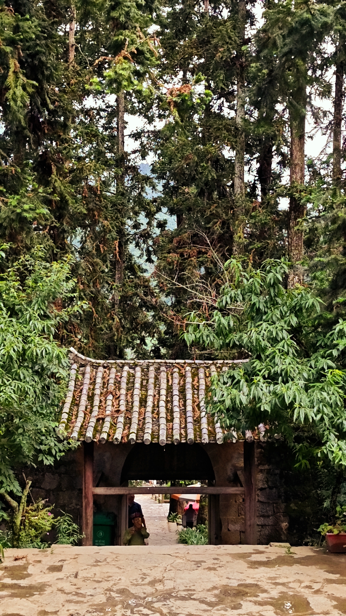 A person sits in a doorway in Sà Phìn Commune, Vietnam. The doorway has a tiled roof and stone walls, surrounded by lush greenery and tall trees. Other people can be seen walking through the doorway.