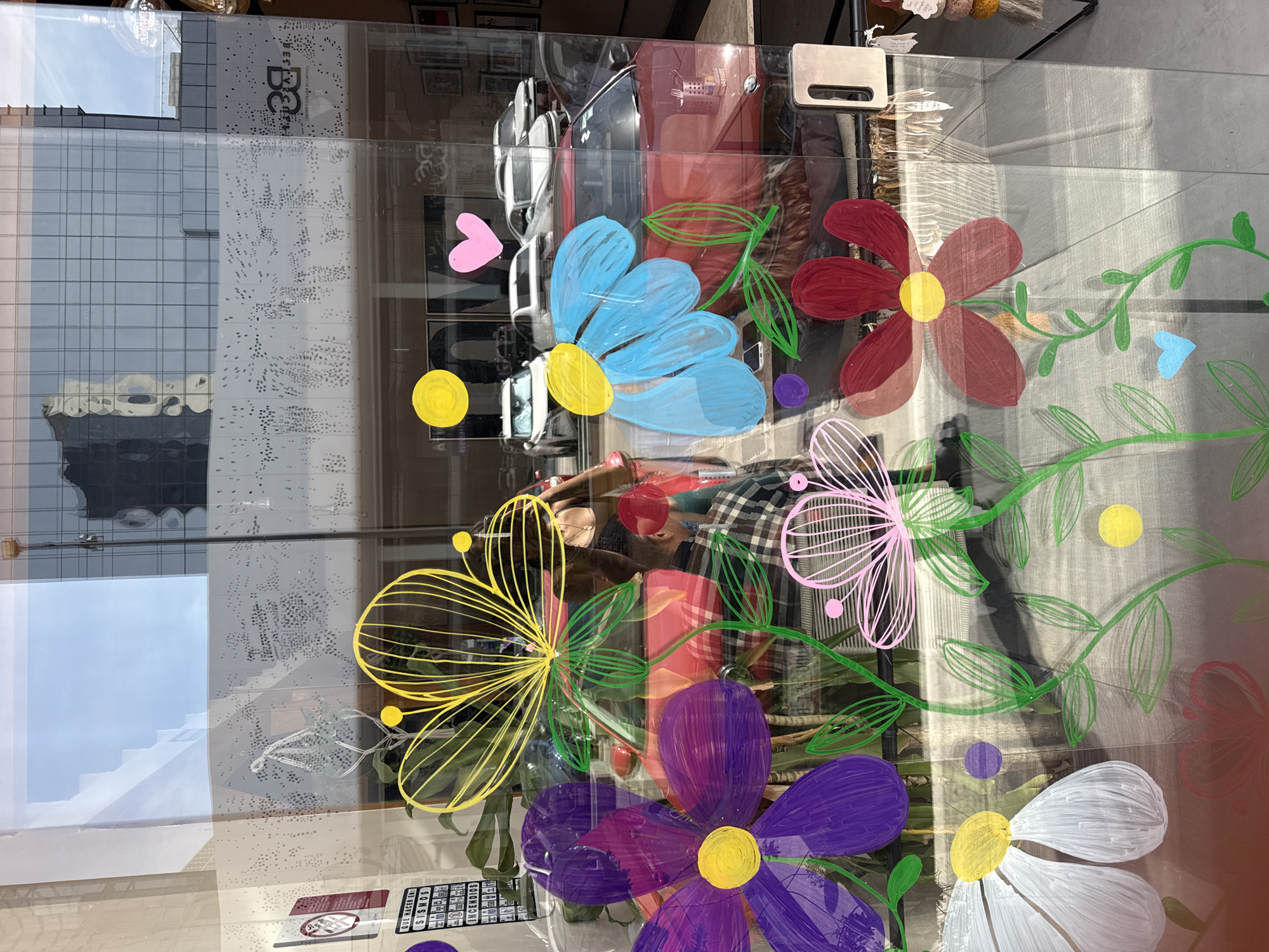 The image shows a window display in Puebla, Mexico, decorated with painted flowers. The flowers are in various colors including blue, red, yellow, pink, purple, and white. Green vines with leaves connect the flowers. Reflections in the glass show a building with white and gray patterned facades, a partially visible car, and interior shop elements. A person is partially visible through the glass. Stickers are adhered near the bottom of the window.