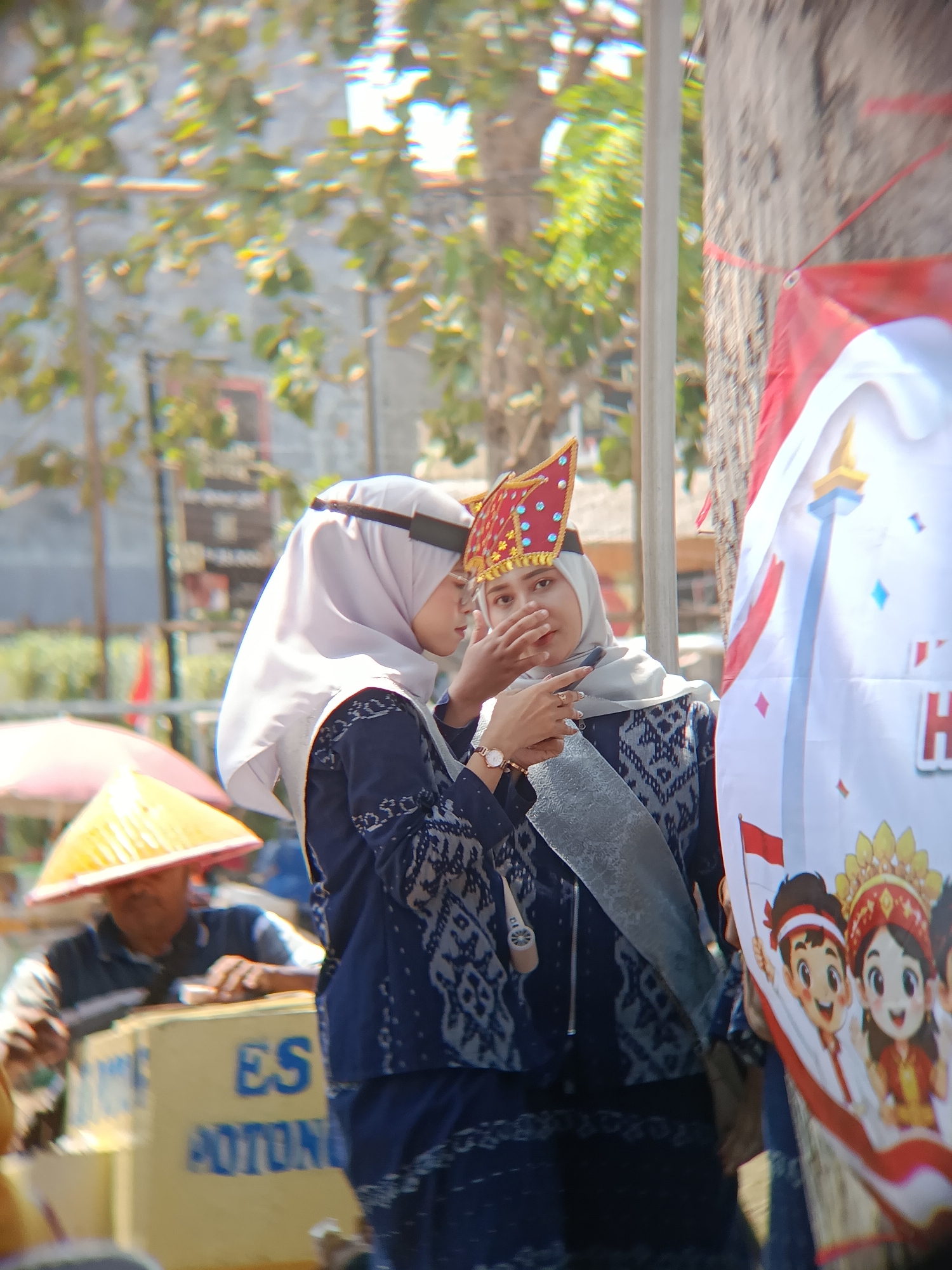 Two young women are standing next to each other outdoors, with trees and buildings in the blurred background. The woman on the left wears a light grey hijab and a dark blue patterned outfit. The woman on the right wears a similar dark blue outfit and a red and gold headdress. She is touching her face with her hand and looking to the side. A banner with cartoon characters and Indonesian flags is visible on the right. In the foreground, a yellow cooler with blue text, partially obscured by a person wearing a conical hat, is visible. The text on the cooler appears to read "ES POTONG," which translates to "ice cream." The scene suggests a local event or celebration in Desa Kebon Manis Kecamatan Cilacap Utara, Indonesia, possibly involving traditional attire.