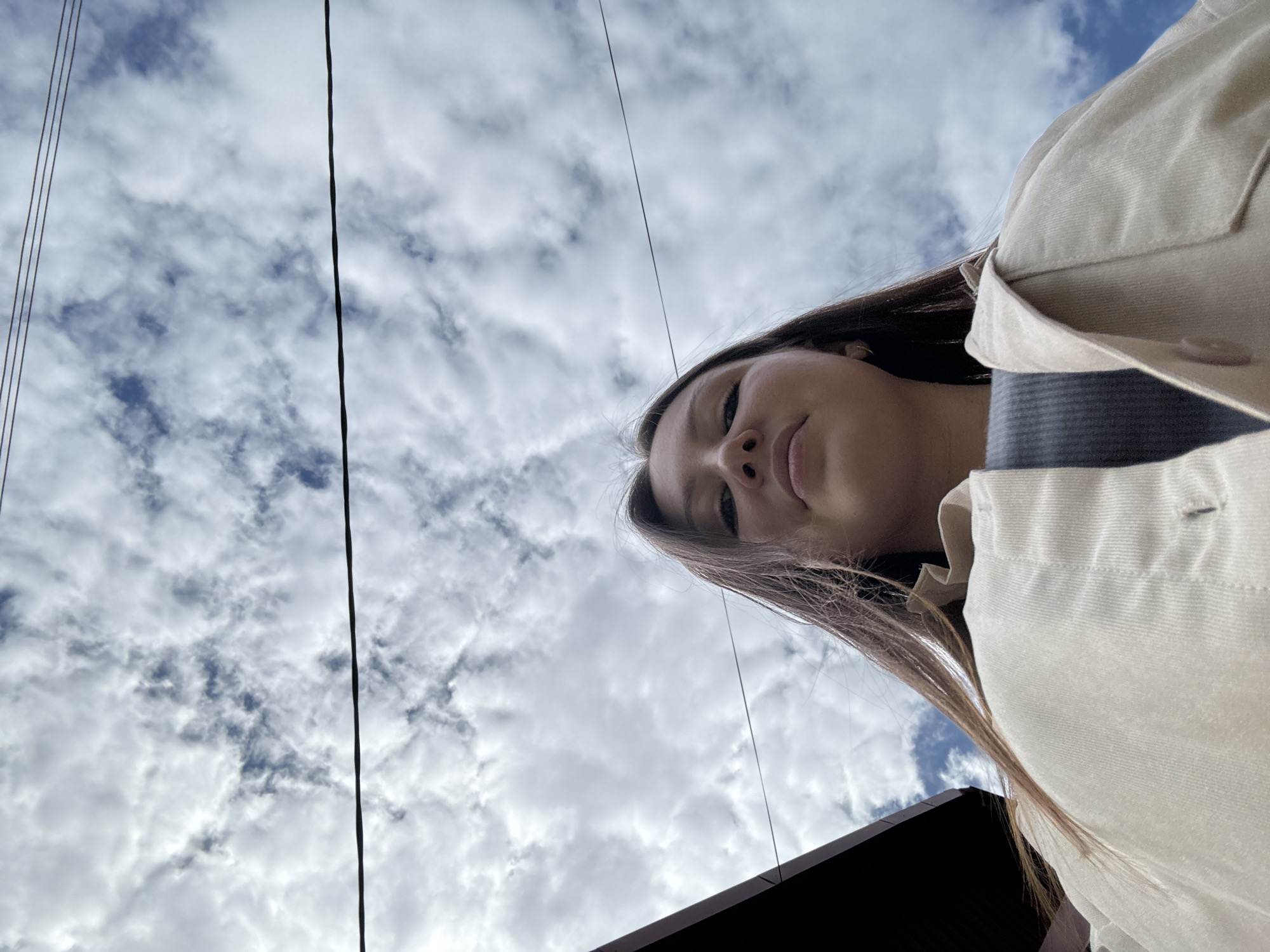 The image is a low-angle, outdoor, close-up selfie of a young woman, seen from the chest up. She has long, brown hair that falls over her shoulders and is wearing a light-colored jacket over a dark grey top. Her face is partially visible, looking up towards the camera. The background is a sky filled with white, fluffy clouds and patches of blue sky. Several thin, dark wires are visible crisscrossing the sky. A dark, angled structure, possibly a roof or building, is visible in the lower right corner of the frame. The lighting suggests it is daytime, and the cloudy sky indicates overcast or partly cloudy weather. The overall mood is casual and personal.