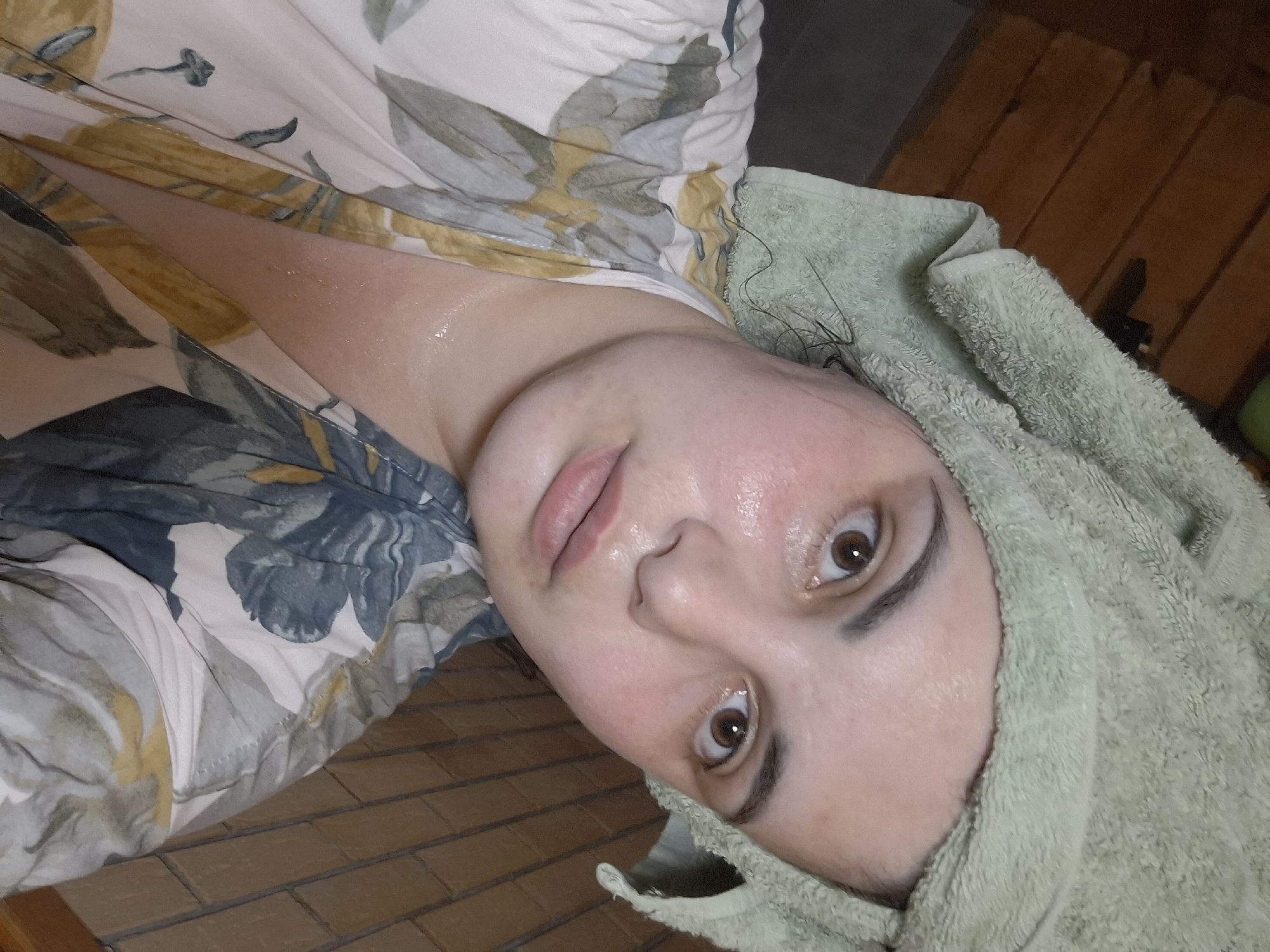 A close-up, tilted selfie of a person with a fair complexion and light brown eyes. The person has a green towel wrapped around their head. Their skin appears slightly flushed and possibly damp. They are wearing a robe or top with a floral print, including shades of green, blue, and yellow. The background shows a wooden floor or wall paneling. The location is unknown and cannot be determined from the image. Амзя, Russia cannot be identified as the city within this image.