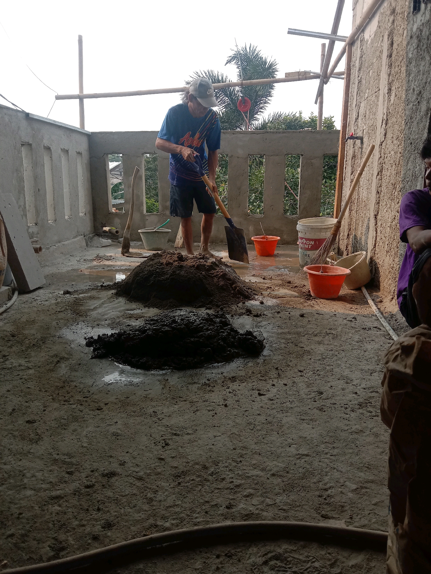 The image depicts a construction site in Tajurhalang, Indonesia, where a man is mixing cement. He is wearing a blue t-shirt, black shorts, and a grey baseball cap. He is holding a shovel and appears to be in the process of combining sand and cement. In the foreground, there are two piles of sand and mixed cement. Several orange buckets, a white bucket, a broom, and a grey bucket are visible near the piles. The ground is covered with concrete and a small amount of water. The background shows a partially constructed building with concrete walls and openings, along with some greenery and bamboo scaffolding. Another person is partially visible on the right side of the image, wearing a purple shirt. The scene appears to be outdoors, possibly on a rooftop or elevated platform.