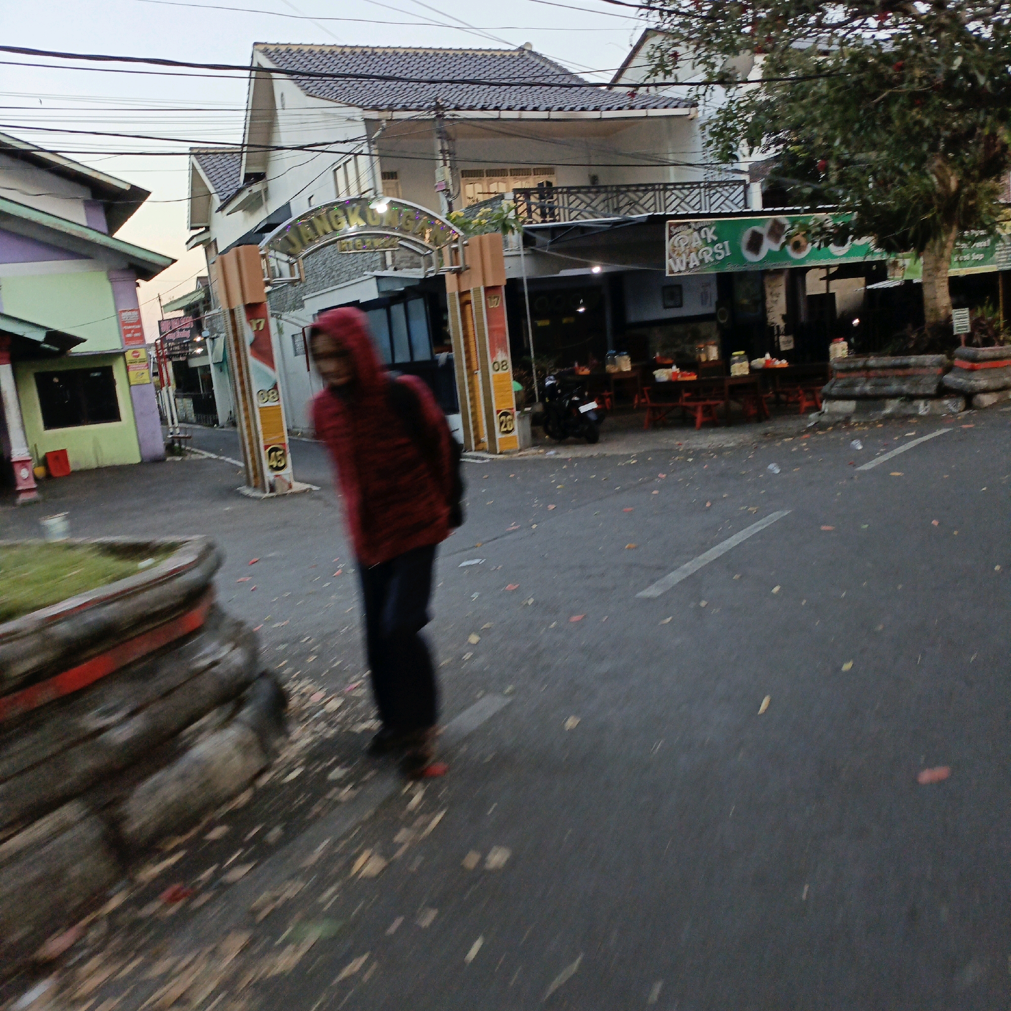 A blurred, medium-shot, eye-level, outdoor, daytime photograph captures a person wearing a red hooded jacket walking on a paved road in Salatiga, Indonesia. The person, facing away from the camera and towards the right of the frame, is mid-stride and appears to be in motion, indicated by the blur. They are wearing dark pants.
Behind the person, a building with a grey tiled roof and white walls is visible, with an arched gateway that has illuminated text reading "JANGKRIK". The gateway is flanked by vertical orange and white structures, one of which displays the numbers "17", "08", and "45". To the right of this building, a green sign with white text, reading "PAK WARSI", hangs above an establishment with outdoor seating and red tables. A tree with green foliage and some red fruits is situated in front of the building to the right.
To the left of the frame, another building with a purple and green facade and a dark window is partially visible. A red cylindrical object stands near this building. The foreground features a dark grey paved road with scattered fallen leaves and a large, textured planter box with red trim. Power lines are strung across the upper portion of the image. The sky is overcast.