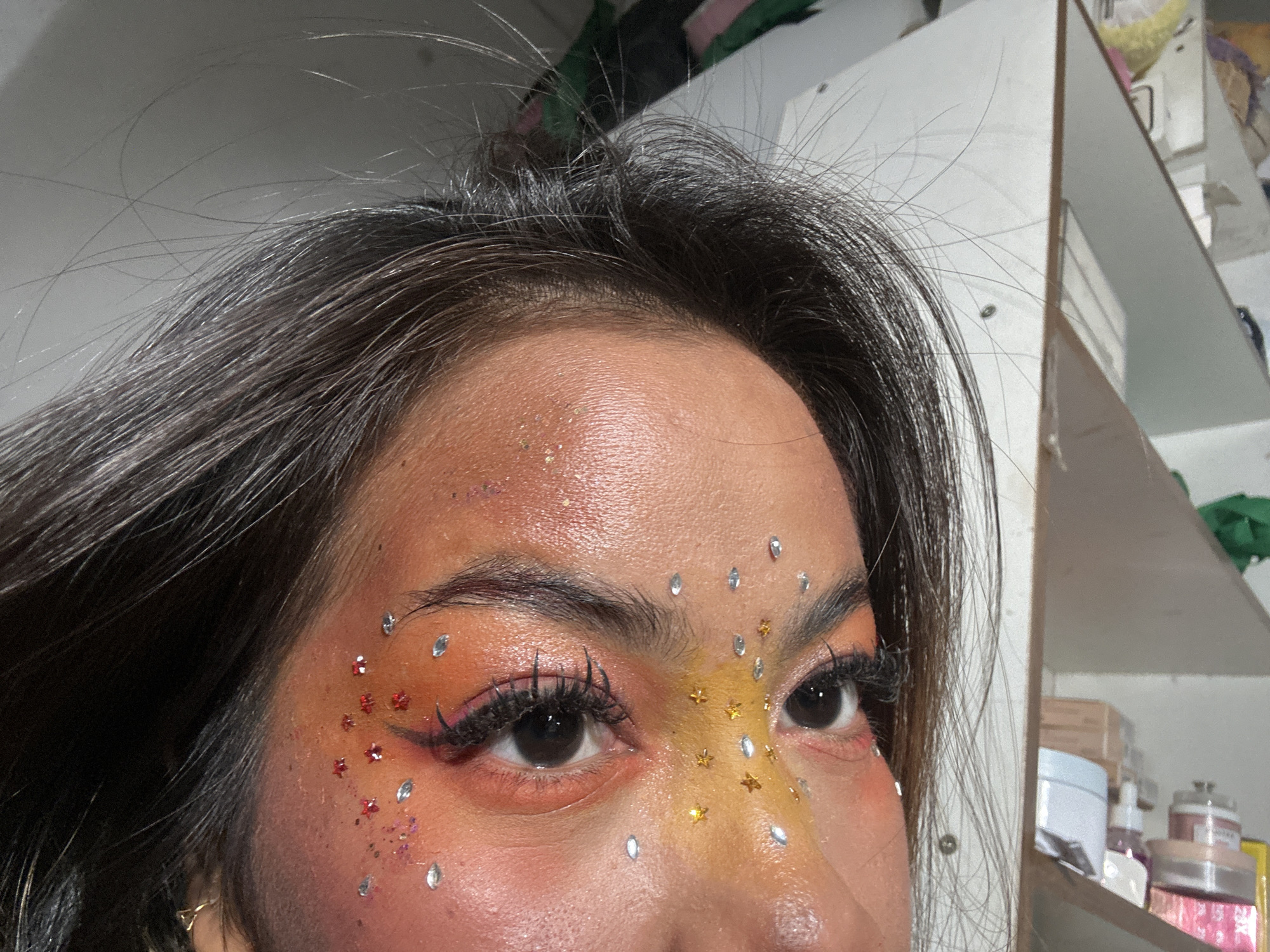 A close-up image focuses on a person's face, specifically the forehead and eye area. The individual has dark hair with some stray strands. The skin appears to have an orange-toned makeup application, with embellishments such as glitter on the forehead and decorative gems near the eyes and cheek. The gems come in two distinct shapes, one being a silver teardrop shape and the other a red star shape. The eye makeup includes defined eyebrows and false eyelashes. The person's gaze is directed slightly to the left.
In the blurred background, there is a glimpse of a white shelf or cabinet filled with various items, including boxes and bottles.
There is no information provided to determine if this image was taken in the city of North Jakarta, Indonesia.