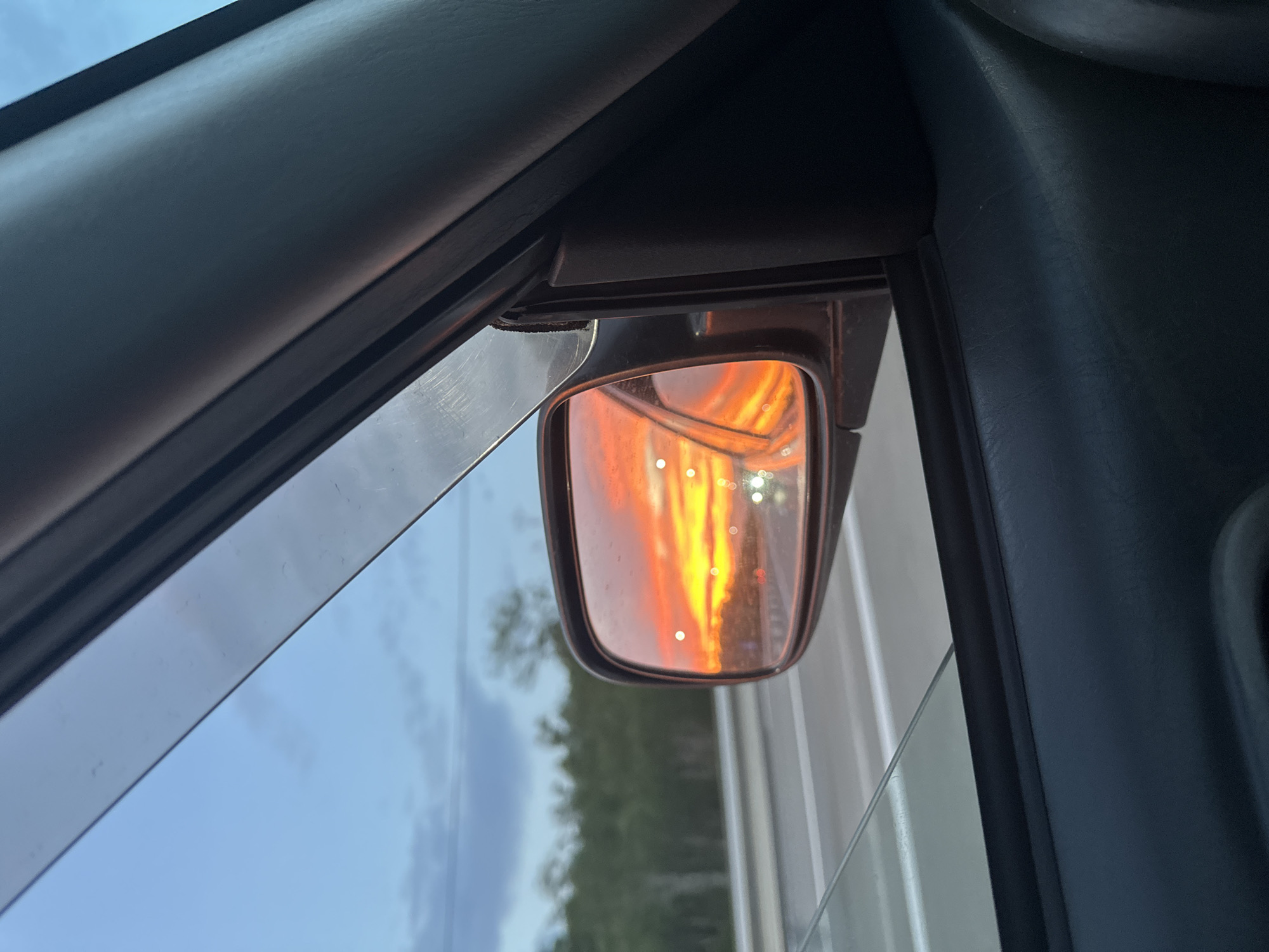 The image shows a view from inside a car, looking out the side window at a sunset reflected in the side mirror. The sky is a vibrant mix of orange, yellow, and pink, indicating a sunset. In the distance, through the side window, trees and a road are visible. The car's interior, including part of the door panel and window trim, is also visible. The reflection in the side mirror is the primary focus, showing the colorful sky and lights from other vehicles on the road. The scene appears to be captured while the car is in motion, as suggested by the blur in the background and the perspective of the reflection. There is no indication of Yelizovo, Russia in the image.