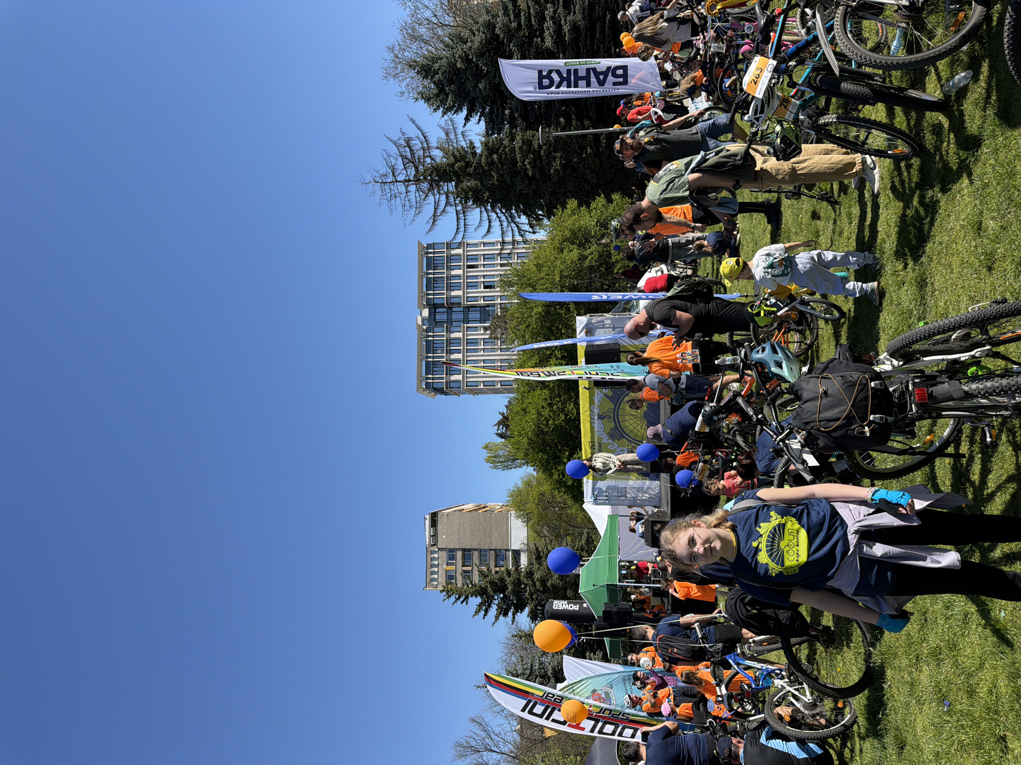Here is a forensic description of the image:
**Scene:**
The image captures an outdoor event in Sofia, Bulgaria, likely a cycling competition or festival. It is daytime with clear blue skies. The setting appears to be a park or grassy area within the city. Modern buildings are visible in the background.
**People:**
Many people are present, mostly adults, with some children visible. They are dressed in casual or athletic attire. Some individuals are wearing orange shirts, suggesting they may be participants or event staff. There is a young girl in the foreground who is wearing a blue shirt with a bike wheel design on it.
**Objects:**
Several bicycles are visible, clustered together. Flags and banners with Cyrillic text, likely advertising the event or sponsors, are prominent. Balloons in blue and orange colors are also present.
**Actions and Interactions:**
There is a gathering of people, seemingly associated with the cycling event. It is not possible to determine specific actions beyond assembly.
**Environment:**
The grassy terrain indicates a park setting. Trees and buildings provide a backdrop, characteristic of Sofia's urban landscape.
**Key Identifiers:**
* Flags and signage in Cyrillic script, indicating the event is held in Bulgaria.
* The Cyrillic word "БАНКЯ" on one of the flags, suggesting a connection to the Bankya region near Sofia.
* The Cyrillic word "СОФИЯ", on the young girl's shirt, which means "Sofia".