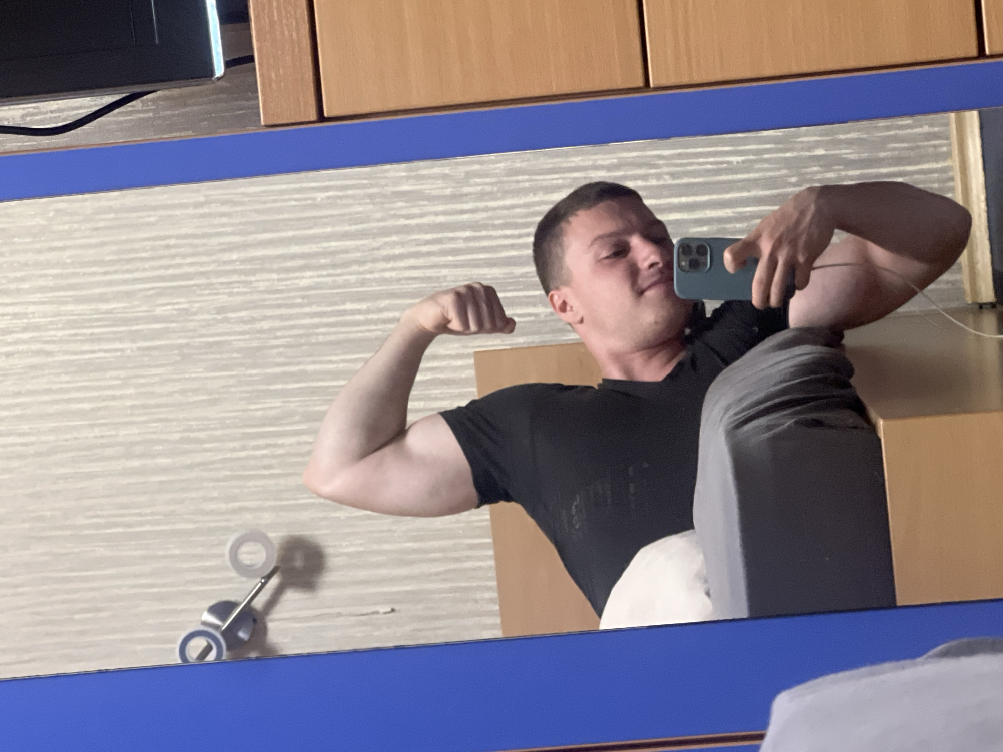 A man is taking a selfie in a mirror, flexing his arm to display his bicep. He is wearing a black t-shirt and grey sweatpants. The mirror is reflecting a room with light brown wooden furniture and striped beige wallpaper. A cable and a television are visible above the mirror. There is a metal object with white and blue rings attached to the lower left corner of the mirror. There is no visual indication of the city of Burgas, Bulgaria.