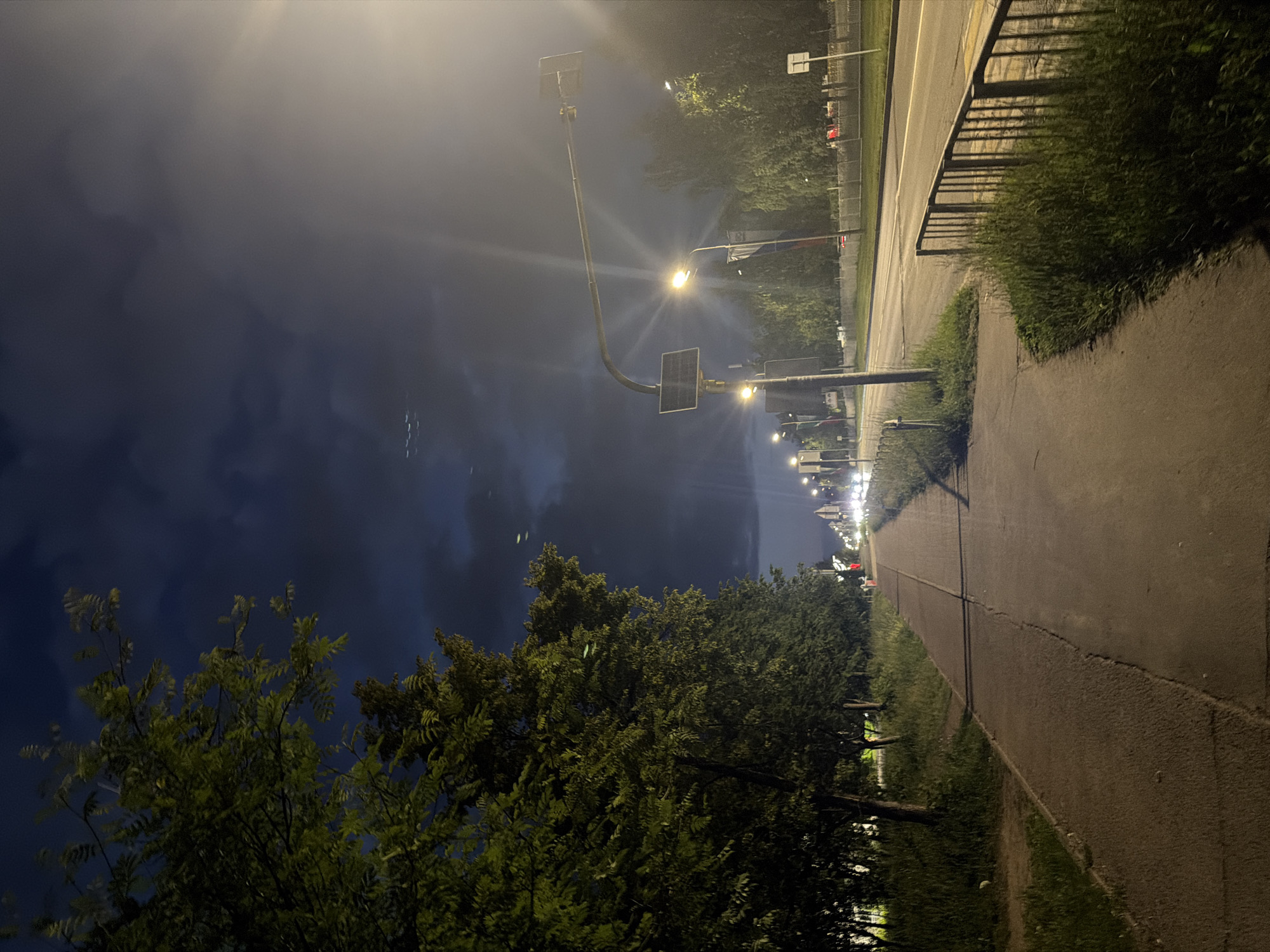 The image depicts a nighttime scene on a sidewalk next to a road in Naberezhnye Chelny, Russia. A row of trees with lush green foliage lines the left side of the sidewalk, casting shadows on the pathway. The sidewalk itself is paved and shows some wear and tear, with a distinct crack running along its length. To the right of the sidewalk is a road with one lane of traffic visible, illuminated by streetlights. Several vehicles can be seen in the distance, their headlights creating streaks of light. A metal fence with vertical bars runs along the edge of the road, separating it from a grassy area. A lamppost with a solar panel is positioned on the sidewalk, providing light. The sky is dark and cloudy, with some patches of lighter sky visible. There are no people clearly visible in the image, but the presence of a road and sidewalk suggests an urban or suburban environment. The overall impression is one of a quiet, possibly deserted, street scene at night.