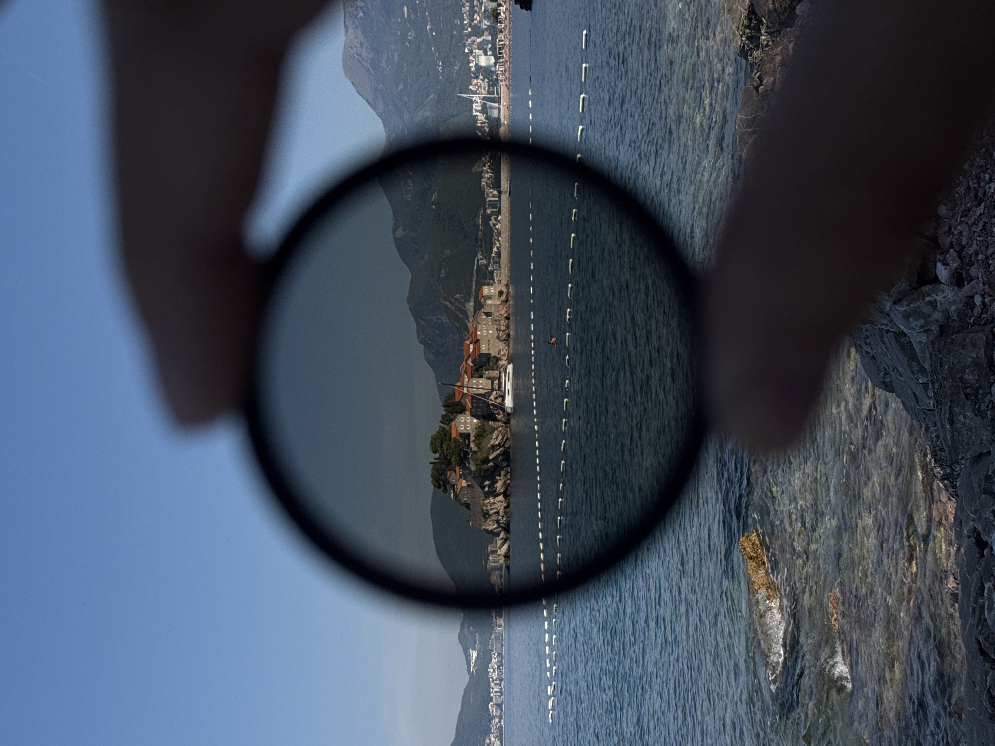 A circular, dark filter is held in the foreground, obscuring a portion of the background scene. Through the clear part of the filter, a sunny daytime view of the sea and a coastal island settlement is visible. The island, identified as Sveti Stefan in Montenegro, features stone buildings with red-tiled roofs, situated on a rocky outcrop. A white sailboat is moored near the island. The sea is a deep blue, with a rocky shoreline in the immediate foreground. The sky above is clear and blue. The filter creates a vignetting effect, darkening the image within its circular boundary, and also casts shadows of fingers holding it onto the foreground.