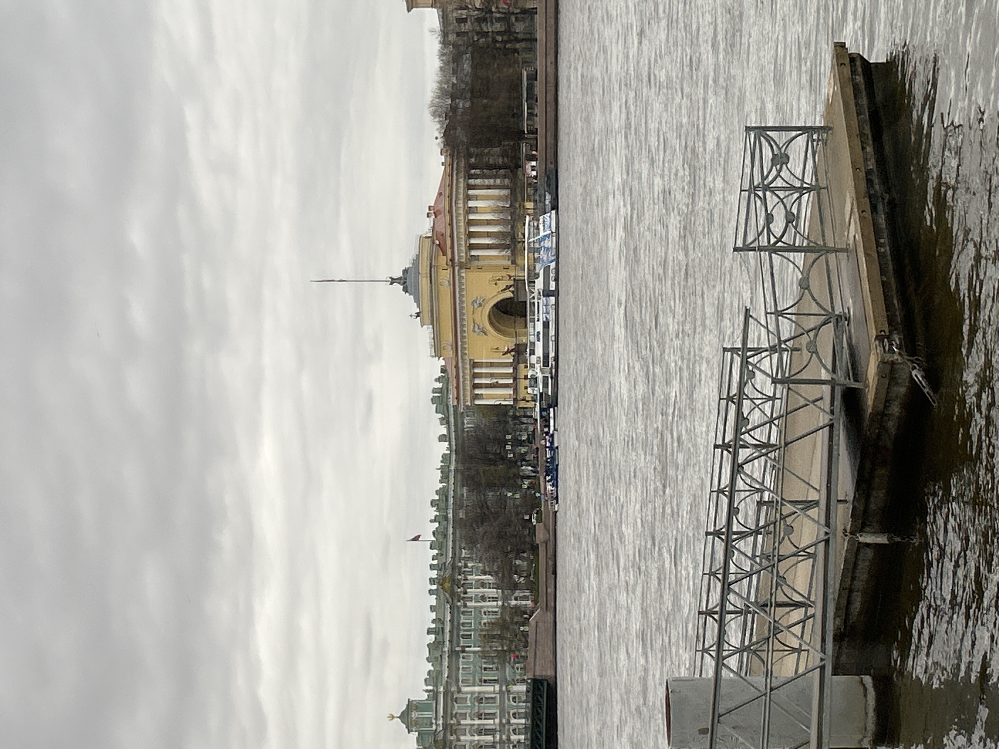 This image captures a panoramic view of a city embankment across a wide body of water under an overcast sky. In the foreground, a metal pier or floating dock with ornate, dark grey railings extends into the water from the right side of the frame. The water, likely a river or canal, appears calm and grey, filling the lower and mid-ground of the scene.
On the far bank, the distinctive architecture of Saint Petersburg, Russia, is visible. A prominent yellow building with classical columns and a tall, slender spire crowned with a ship weather vane stands as a central landmark, identifiable as the Admiralty. To its left, a lengthy complex of grand, multi-storied buildings, painted in shades of light green and blue, lines the embankment, recognized as part of the Winter Palace and Hermitage Museum complex. These buildings feature numerous windows and intricate detailing. Along the embankment, bare trees with no visible foliage are scattered, suggesting a cooler season. Several white boats or tour vessels are moored near the waterfront in front of the yellow building.
The sky is uniformly light grey and overcast, indicating a dull, sunless day. The overall lighting is soft and diffused. The scene is static, devoid of any visible human activity or movement. The cool color palette and absence of foliage contribute to a tranquil, somewhat muted atmosphere. No discernible text is visible within the image.