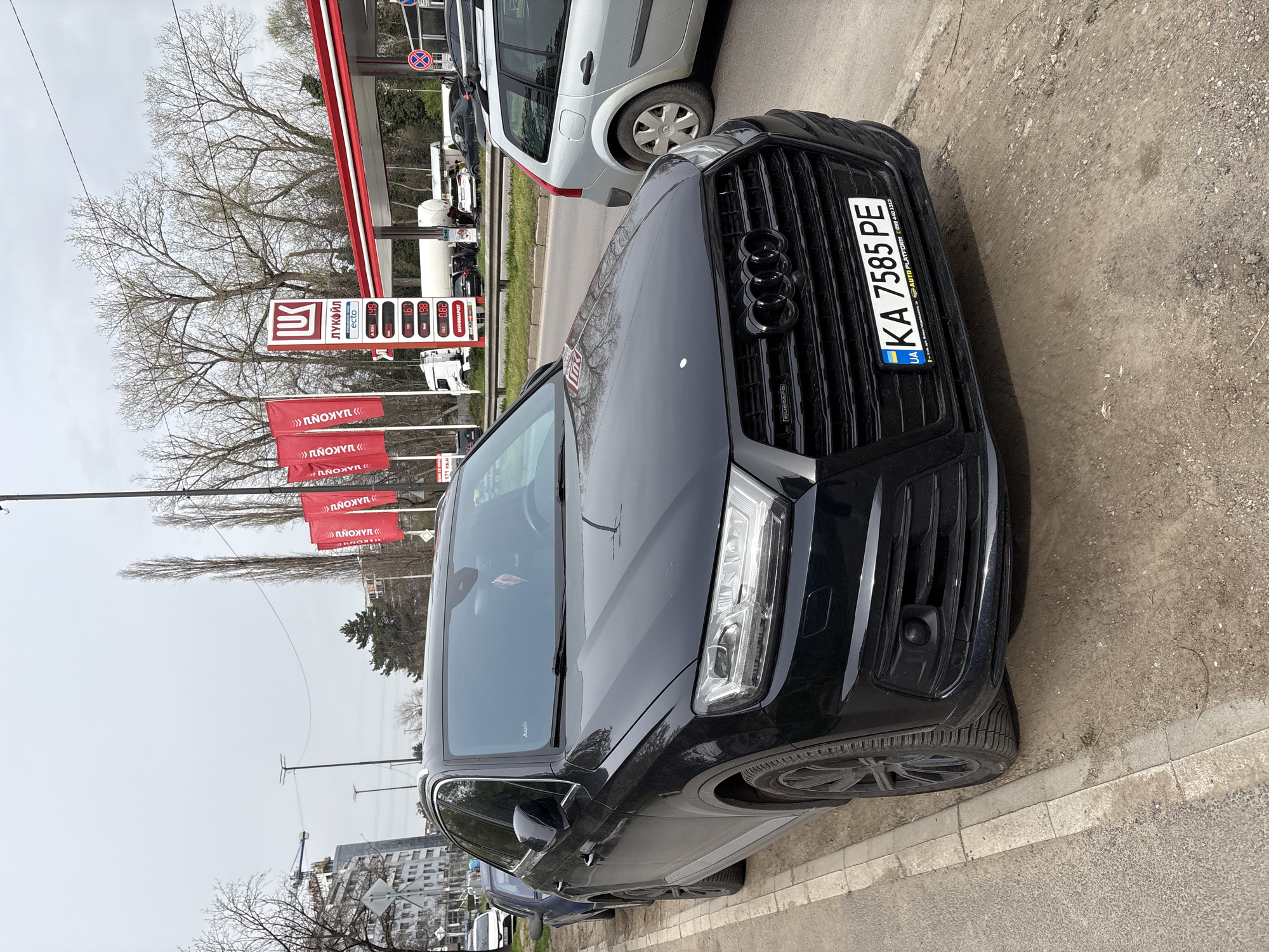 The image shows a black Audi Q7 parked on the side of the road. The car has a license plate with the number "KA 7585 PE". The license plate also indicates that the car is registered in Ukraine. Behind the car is a Lukoil gas station, recognizable by its logo and red flags with Cyrillic writing. The gas prices are displayed as well. Based on these details and other signage visible, the location is likely in Sofia, Bulgaria. Other vehicles are also present on the street. The weather appears overcast. The car is parked next to a concrete curb.