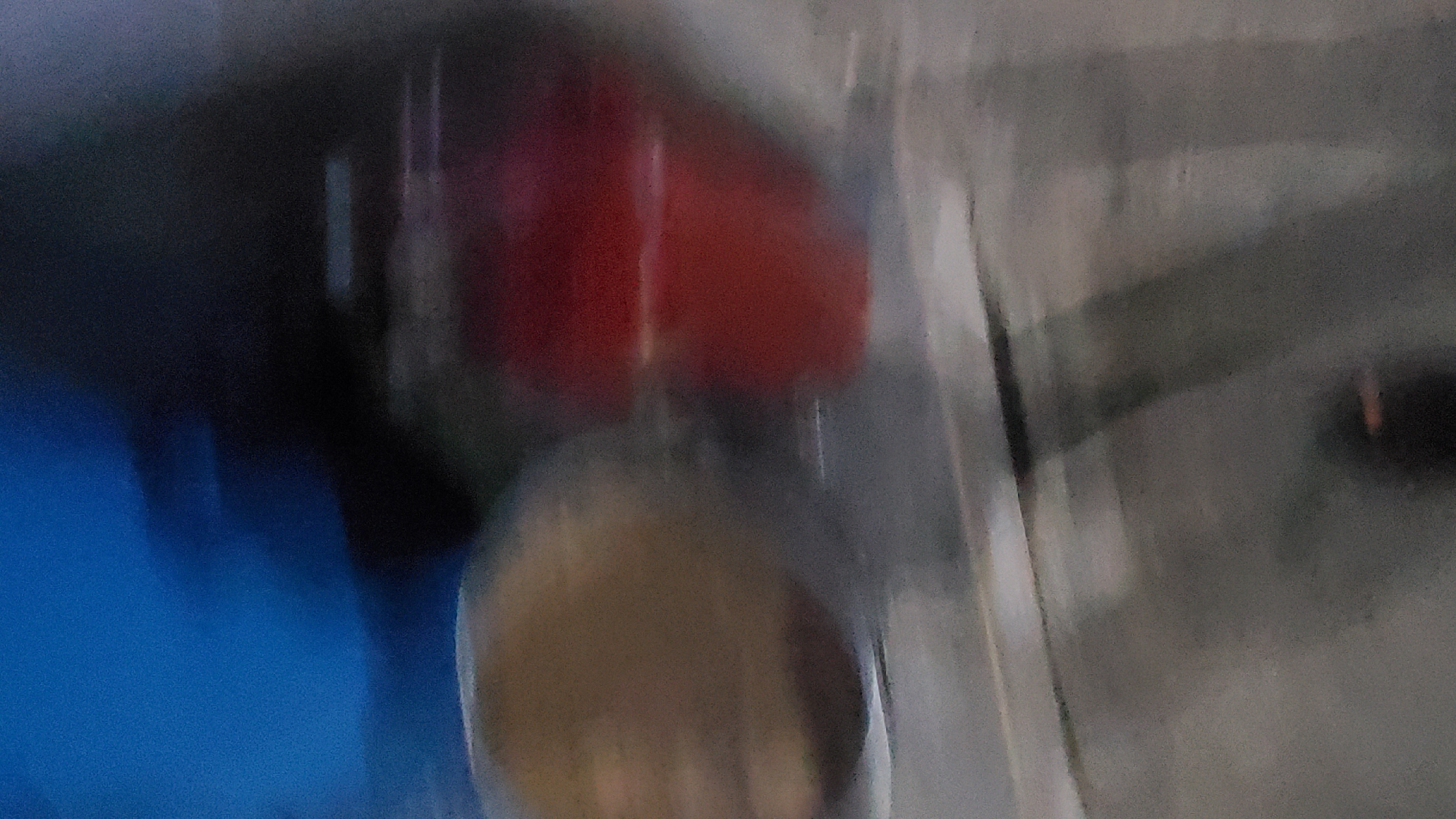 This image, captured in the city of Jalingo, Nigeria, presents a highly blurred and indistinct scene, indicative of significant motion or focus issues. The upper left quadrant features a large, amorphous area of bright blue, transitioning to a dominant dark, almost black region across the upper and central right portions of the frame. In the mid-left, a blurred, rounded object with a tan-brown hue is partially discernible. To its immediate right, a prominent, vertically oriented, block-like object in vibrant red is visible, containing subtle horizontal striations. The lower third of the image consists of a grey-toned surface exhibiting pronounced horizontal streaking, suggestive of pavement or a similar ground plane. No distinct human figures, specific actions, or detailed environmental elements are identifiable within the extensive blur.