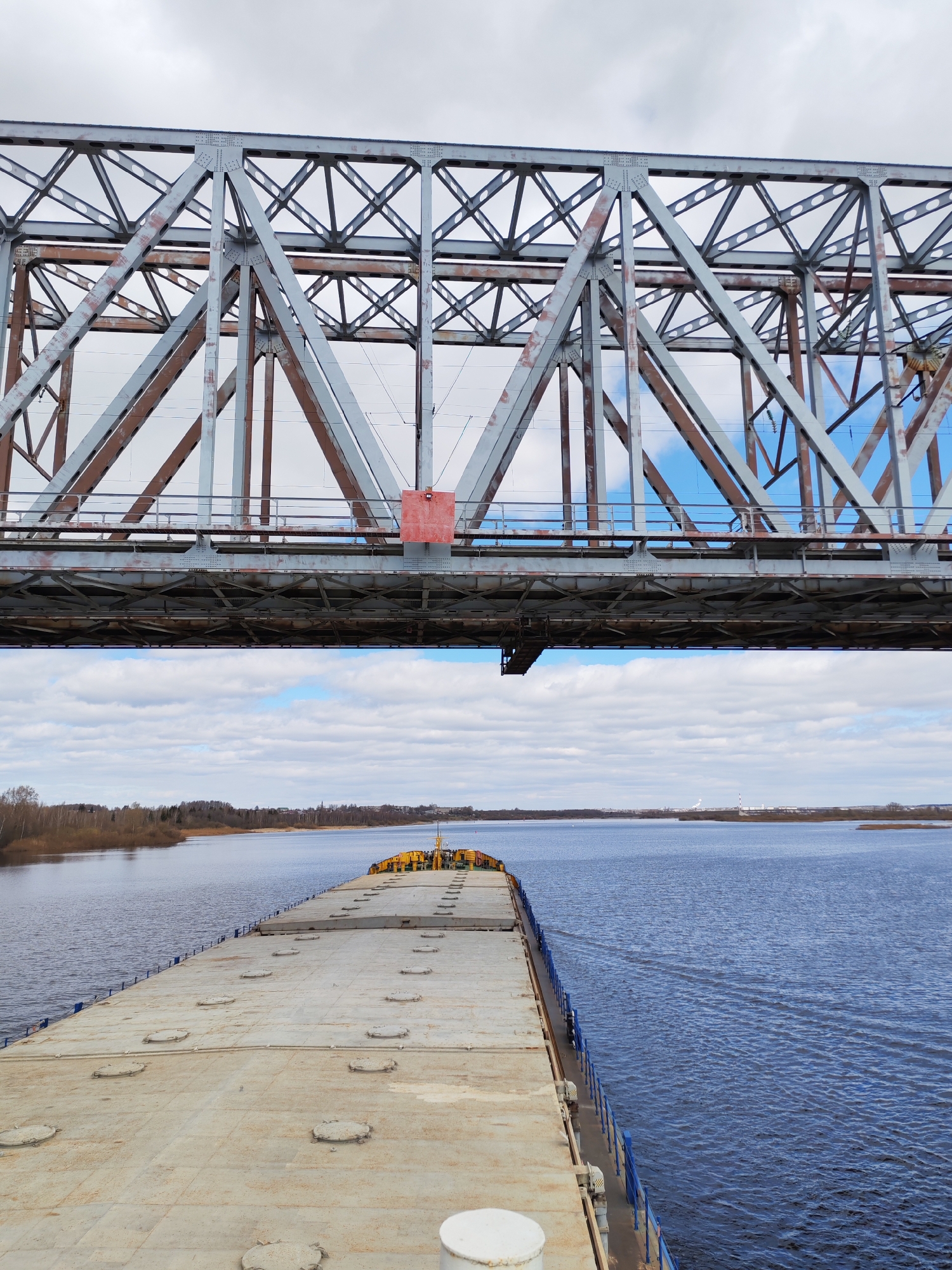 A barge navigates a river beneath a steel truss bridge. The barge has a flat, concrete-like surface with circular indentations and is bordered by blue railings. On the barge's stern, there is yellow equipment. The river stretches into the distance, with land visible on both sides. In the background, faint city structures are visible, along with overcast skies. The bridge's structure is metal, with a reddish-brown hue on the exposed sections.