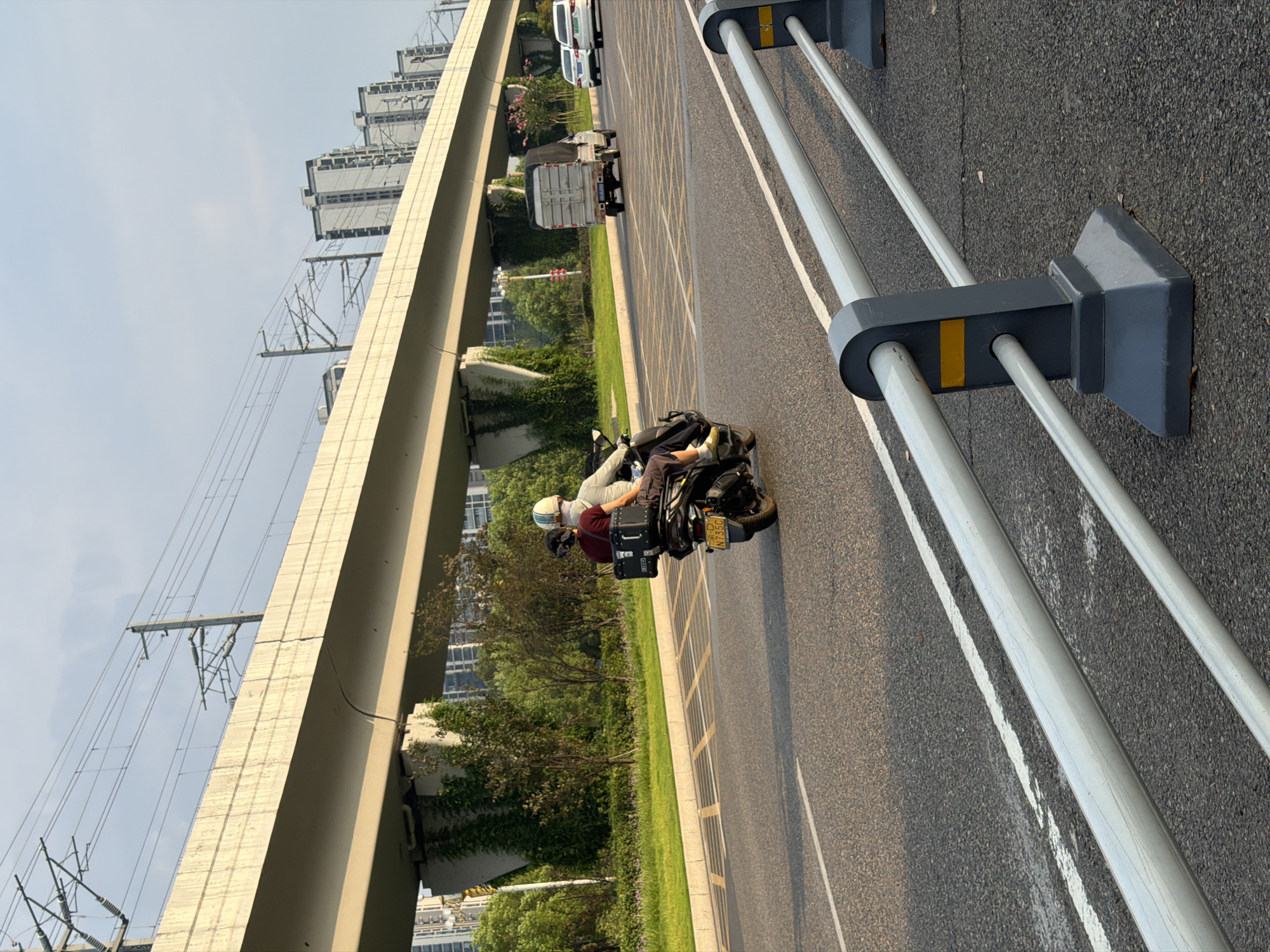 The image captures a street scene in Jiangjin, China, likely during the daytime under a bright sky. In the foreground, a metal guardrail with gray posts and silver bars separates the road from the viewer. The road itself is asphalt with white lane markings.
On the road, a motorcycle is traveling away from the camera. The motorcycle carries two people, both wearing helmets. The person in the rear, dressed in a maroon shirt, has a black storage box mounted behind them. The person in the front is wearing a light-colored jacket. The motorcycle's license plate is visible, displaying "N735C".
Further down the road, a small white truck with a silver cargo box is in motion. Behind the truck, two white cars are also visible, suggesting traffic on the road.
Overhead, a large elevated concrete structure, likely an overpass or a section of an elevated railway, dominates the background. Power lines and their supporting poles are strung across the sky above the overpass. The overpass is supported by substantial concrete pillars, some of which are covered in green ivy.
Beyond the overpass, a row of modern, multi-story residential buildings is visible against the skyline. Lush green trees and vegetation line the side of the road beneath the overpass. The overall environment appears to be an urban setting with significant infrastructure development.