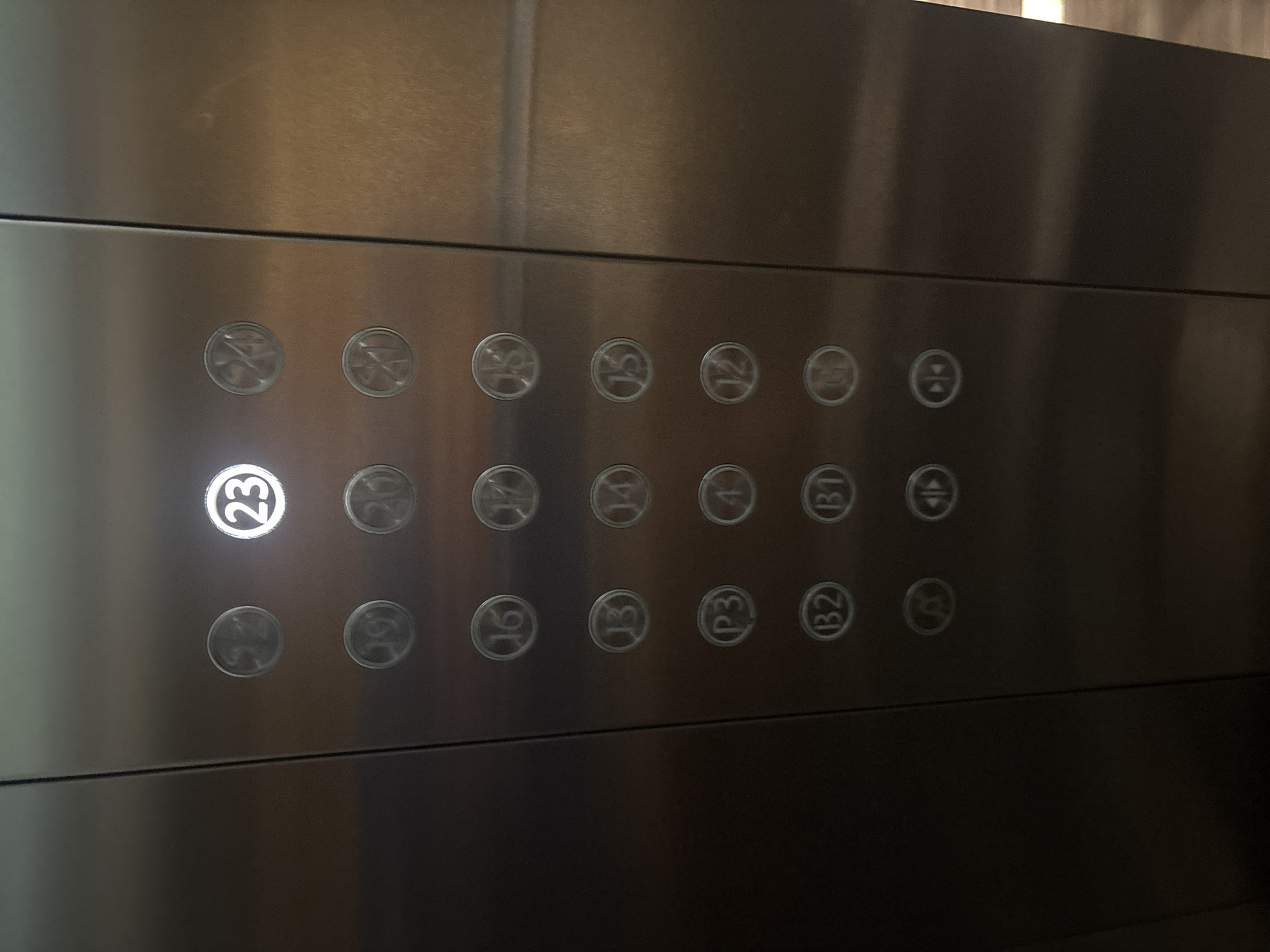 The image shows the elevator control panel of a building. The panel is made of stainless steel and has multiple circular buttons, each with a number or letter inscribed on it. The buttons are arranged in columns and rows. The button labeled "23" is illuminated, indicating that the elevator is currently at the 23rd floor. Other buttons visible include numbers from 12 to 22, as well as buttons labeled "P3", "B2", "B1", "A", and two buttons with arrows (likely for door open/close and floor up/down). The lighting is somewhat dim, and there are reflections on the metal surface. The environment appears to be an indoor space, likely the interior of an elevator. There is no specific indication of the city of Dubai, United Arab Emirates, within the image itself.