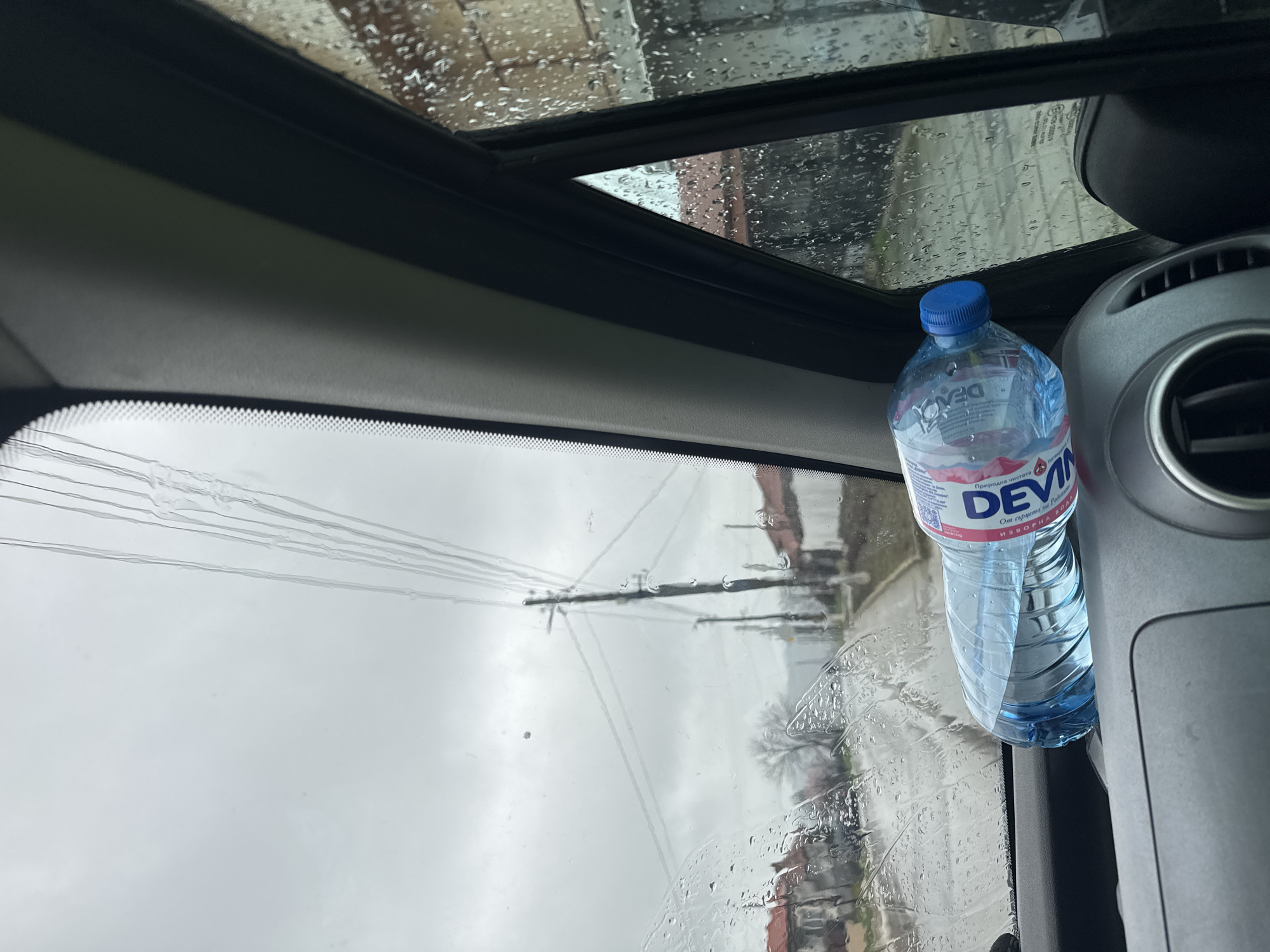 The view from inside a car shows a rainy day in Lyuben, Bulgaria. A bottle of "Devin" brand water sits on the dashboard to the right of the frame. The windshield is covered with raindrops, obscuring the view of buildings and power lines outside. A sunroof is partially visible above the windshield, also covered in rain.