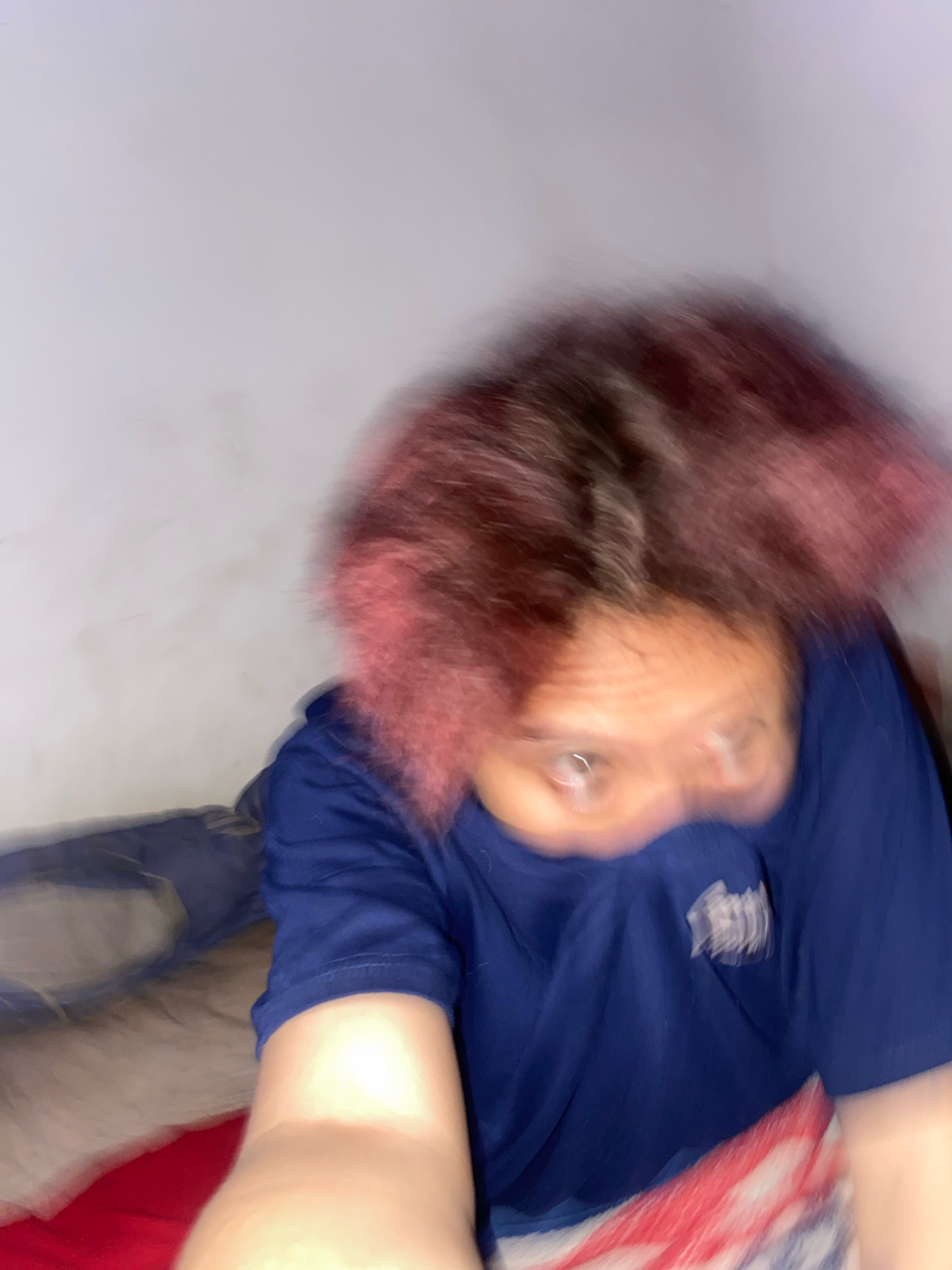 The image shows a close-up of a person's head and upper body, captured with a motion blur that suggests rapid movement. The person has reddish-pink, voluminous hair and is wearing a dark blue t-shirt. Their eyes are visible and appear wide. The background is a plain, light-colored wall, and the lower portion of the image shows bedding with a red and white striped pattern. The context of Klurak, Indonesia, is not discernible from the visual information in the image itself.