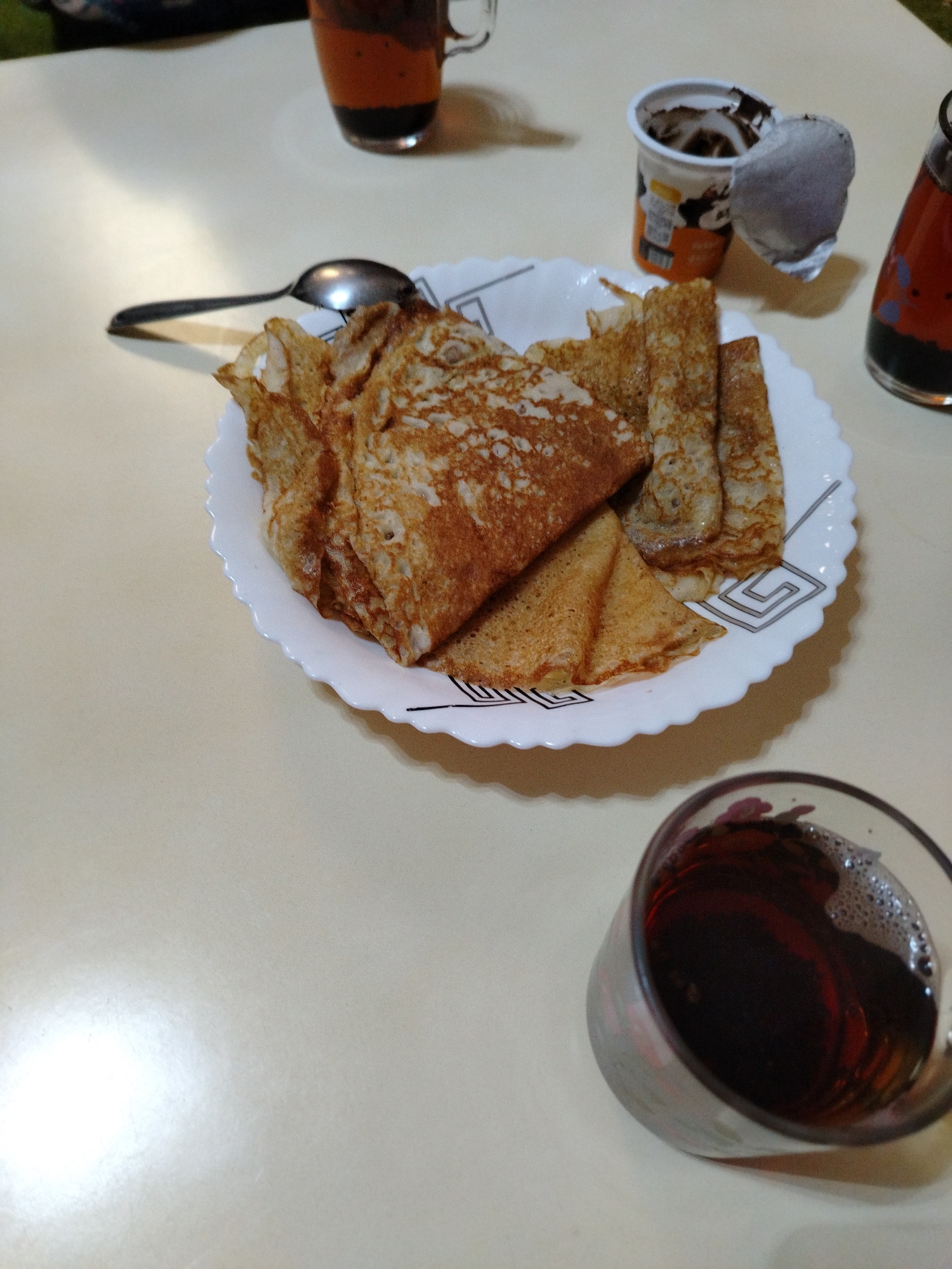 On a light-colored surface, possibly a table, a plate of folded crepes is centered in the image. Adjacent to the plate are a spoon, a yogurt cup with the lid partially peeled back, and two glasses containing dark liquid. One glass appears to be a mug with two layers of liquid; the other is a clear glass with a wider rim. The environment suggests an indoor setting, possibly a home. The precise location, Al Mas'oudi, United Arab Emirates, cannot be verified from visual information.