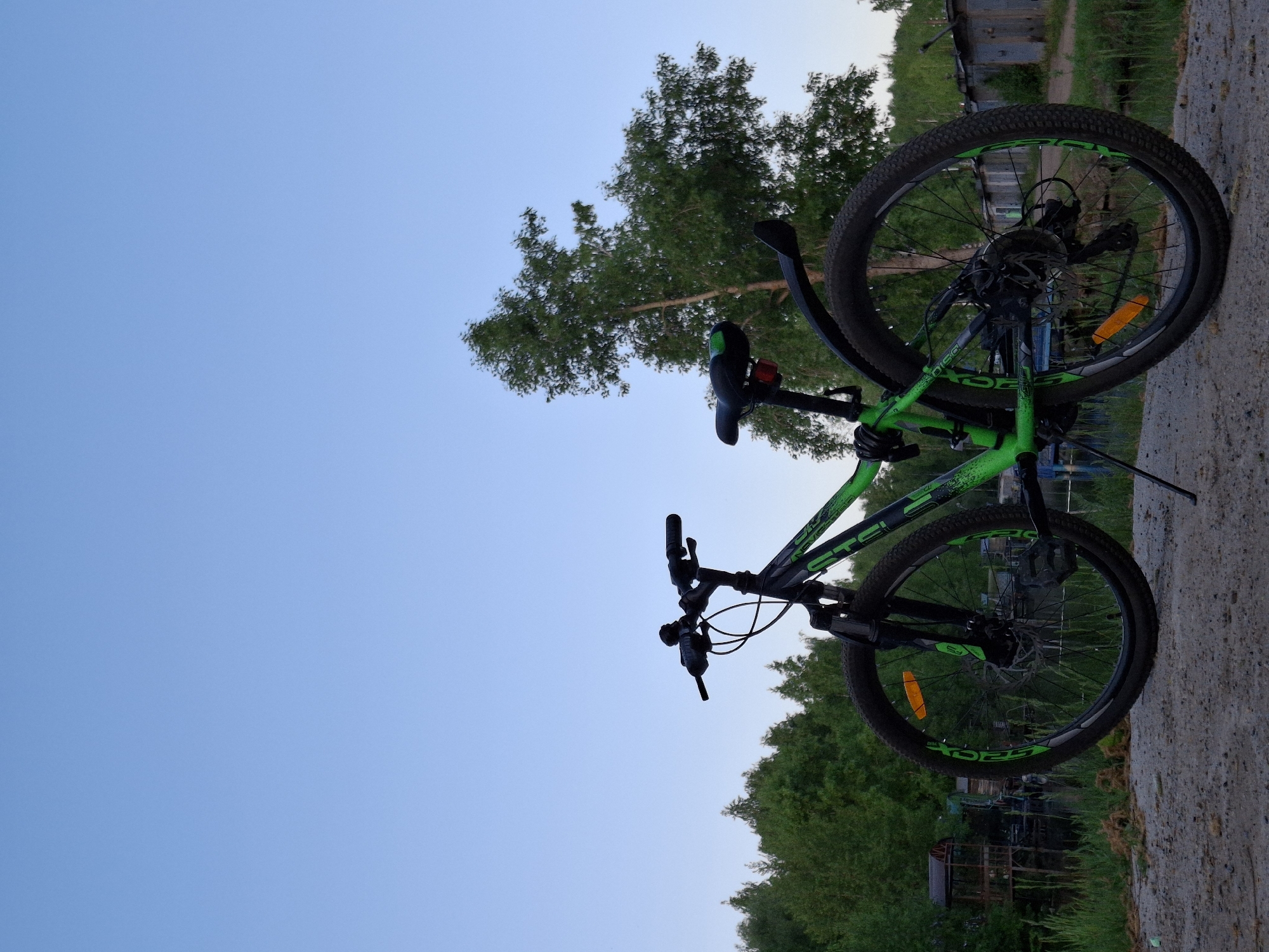 A green and black bicycle is situated on a concrete surface near a water body, likely a river or pond. The background features trees and a few small buildings. The photograph was likely taken in a location near the city of Chaikovsky, Russia. The sky is light blue.