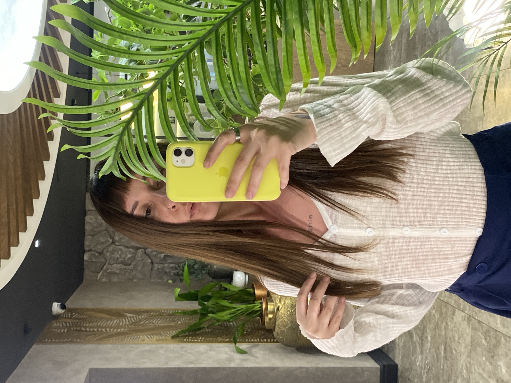 A woman is depicted taking a mirror selfie in an interior setting, identified as being in Votkinsk, Russia. She holds a bright yellow iPhone 11 or 12 in her right hand, partially obscuring her face, which features straight bangs and a visible nose ring. Her left hand runs through her long, straight brown hair. She wears a sheer, ribbed white long-sleeved button-down top and dark blue bottoms, likely a wrap skirt, secured by a single blue button. A silver-toned ring is on her right ring finger, and a partially visible, reversed text tattoo (appearing as 'mirr') is on her upper chest.
The reflected background reveals an elaborately decorated interior. To the left, a decorative golden-toned, vertically patterned screen is on a grey wall, next to an ornate golden pedestal displaying several green potted plants. To the right, a large artificial green fern partially obstructs a grey stone-textured wall. The ceiling, visible in the reflection, features dark grey panels, wooden slats, a white circular light fixture, and a white security camera mounted on a dark panel. The floor is tiled with light-colored, possibly polished stone, squares.