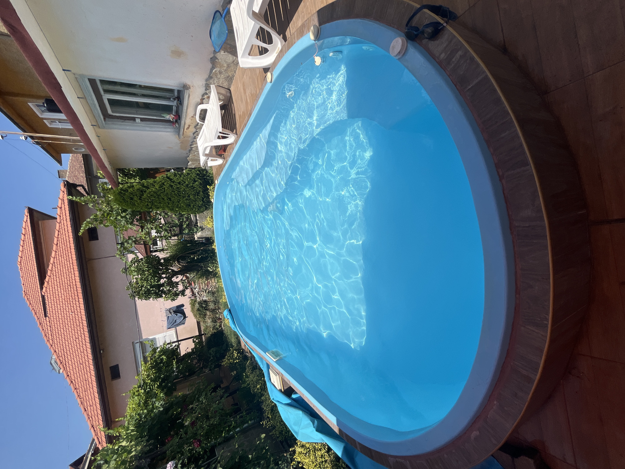 A clear blue swimming pool with a light blue rim is situated on a wooden deck in a sunny backyard. The pool has built-in steps and is partially covered with a blue tarp. Two white lounge chairs and a blue pool skimmer are placed on the deck next to the pool. In the background, there are houses with red-tiled roofs, lush green trees, and bushes. The scene appears to be a residential property in Pavlikeni, Bulgaria, on a bright, clear day.