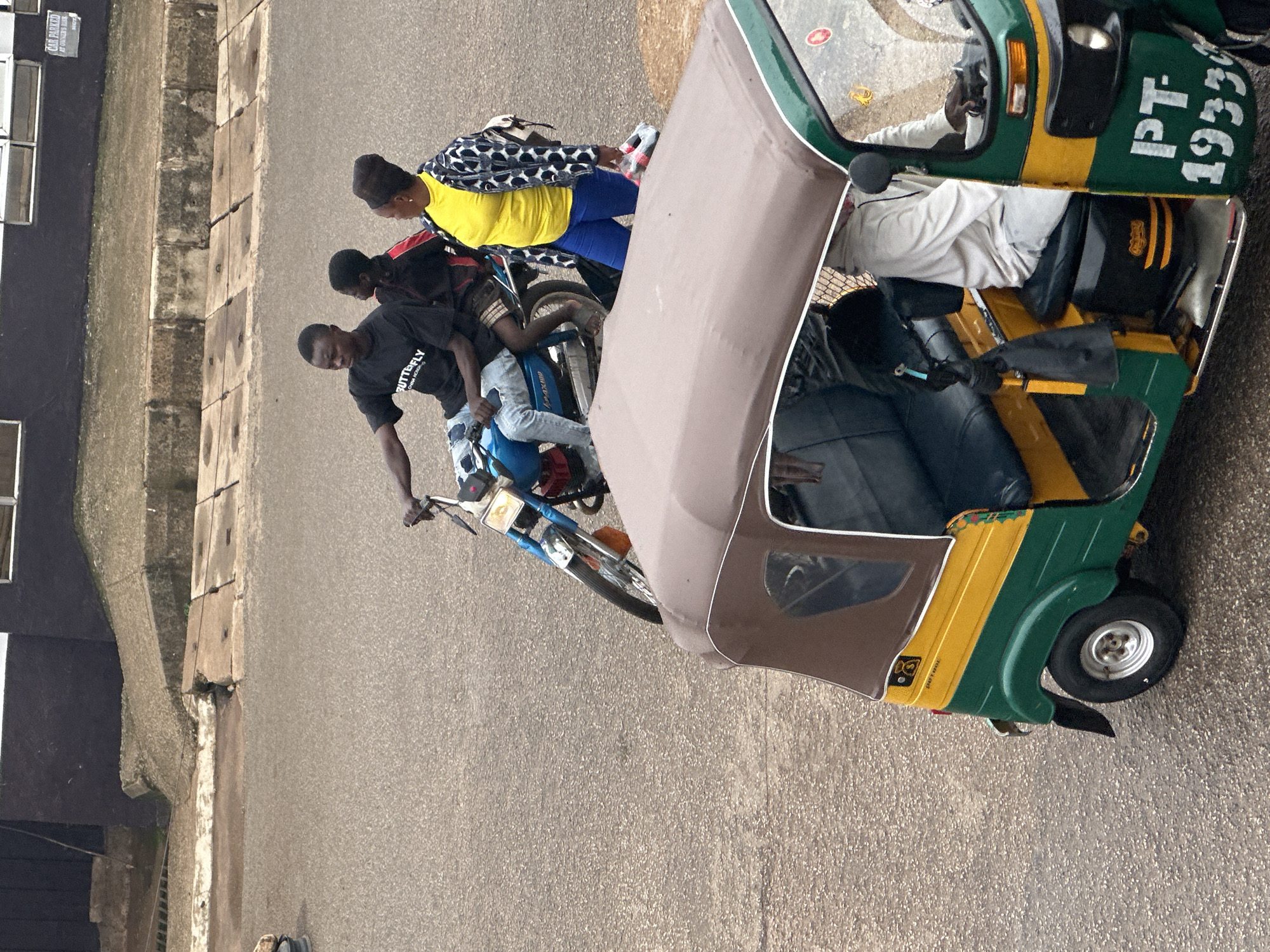This media file depicts a scene in Bukuru, Nigeria, featuring a three-wheeled vehicle, commonly known as a "Keke Napep" or "Maruwa," and several individuals. The Keke Napep is predominantly green and yellow, with visible text on its side reading "PT 1933". It has a fabric canopy over the passenger area.
Four people are associated with the Keke. One individual, wearing a black t-shirt with white text that appears to say "BUTTERFLY", is seated in the driver's position. Another person, in a yellow shirt and blue pants, is seated behind the driver, wearing a patterned jacket. Two more individuals are positioned behind the first two passengers. The second person from the front appears to be wearing a black shirt with red accents, and the person furthest back is mostly obscured. One of the passengers has a helmet resting on their lap.
The Keke itself is a blue motorcycle, integrated into the three-wheeled vehicle. The surrounding environment appears to be a paved road or street, with a concrete structure running alongside it on the left. This structure has several rectangular concrete slabs with dark outlines. On the far left, part of a building with windows is visible.
The scene appears to be taking place during daylight, with the lighting suggesting it could be a bright day. There are no obvious indicators of specific weather conditions. The general atmosphere seems to be one of travel or waiting for transport, with the people positioned as if they are about to embark on or have just completed a journey. The image is shot from a slightly elevated angle, looking down at the scene.