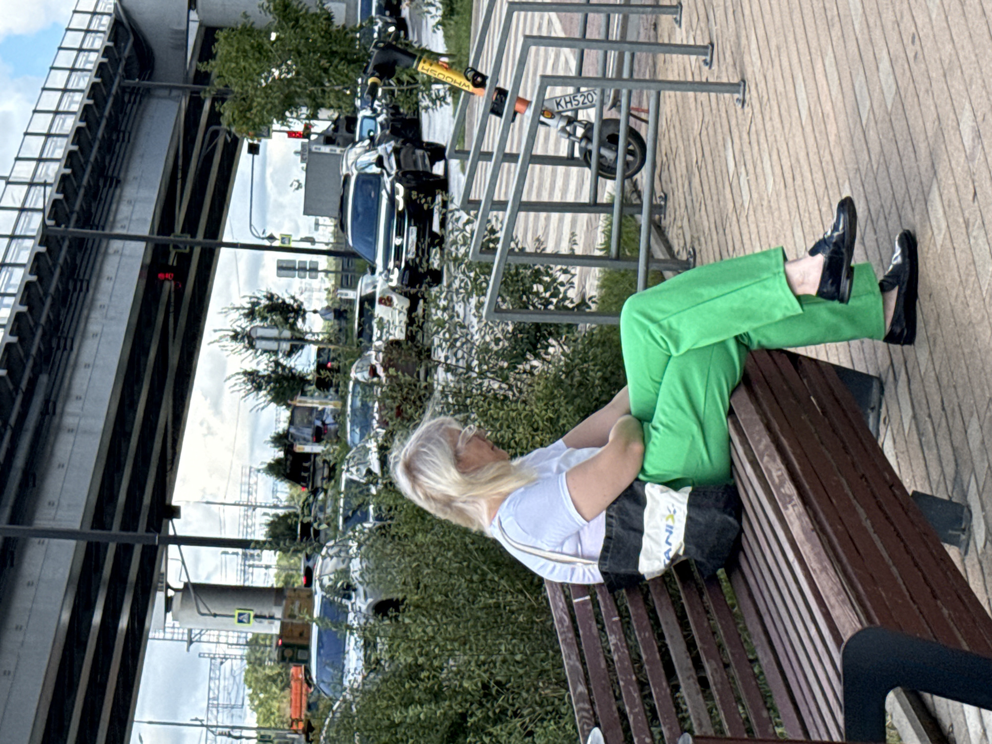 A woman with blonde hair, wearing glasses, a white t-shirt, and bright green pants, sits on a brown wooden bench. She has a black and white tote bag on her lap, with the word "ANIX" visible on it. The bench is positioned on a paved area, possibly a sidewalk, with metal railings to the right of the woman. Beyond the railings, several cars are visible, including a black SUV and other vehicles in traffic. In the background, an elevated highway structure is prominent, with power lines and trees interspersed. The scene appears to be in an urban environment, likely a street or public space within the city of Moscow, Russia, given the style of the vehicles and infrastructure. The lighting suggests it is daytime, with a partly cloudy sky.