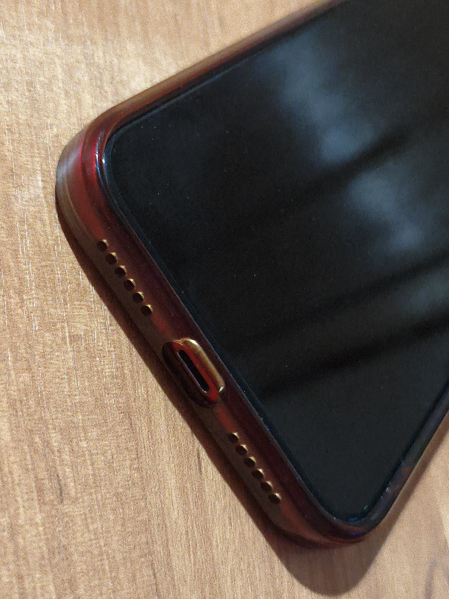 The image shows a close-up, angled view of a smartphone with a dark screen, resting on a light brown, textured surface resembling wood grain. The phone has a reddish-brown, translucent case with a metallic gold accent around the charging port. Six small circular holes, likely speaker grilles, are visible along the bottom edge of the phone, to the left of the charging port. The lighting suggests an indoor environment, with reflections visible on the screen and case. The scene is static with no people or activity depicted. The location in Lyubertsy, Russia is not discernible from the image itself.