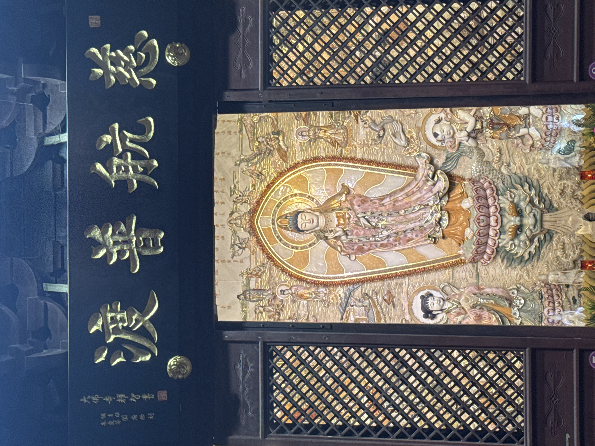 A close-up shot shows an ornate panel in Yuexiu District, China. On a black panel, gold Chinese characters run vertically, accompanied by small, round gold embellishments. To the right, separated by a lattice-patterned screen, a vertical rectangular panel displays a three-dimensional relief sculpture. The sculpture features a central, reclining figure with a halo, adorned in robes, surrounded by various figures and ornate decorations. The entire scene suggests a religious or cultural artifact, likely found in a temple or historical site.