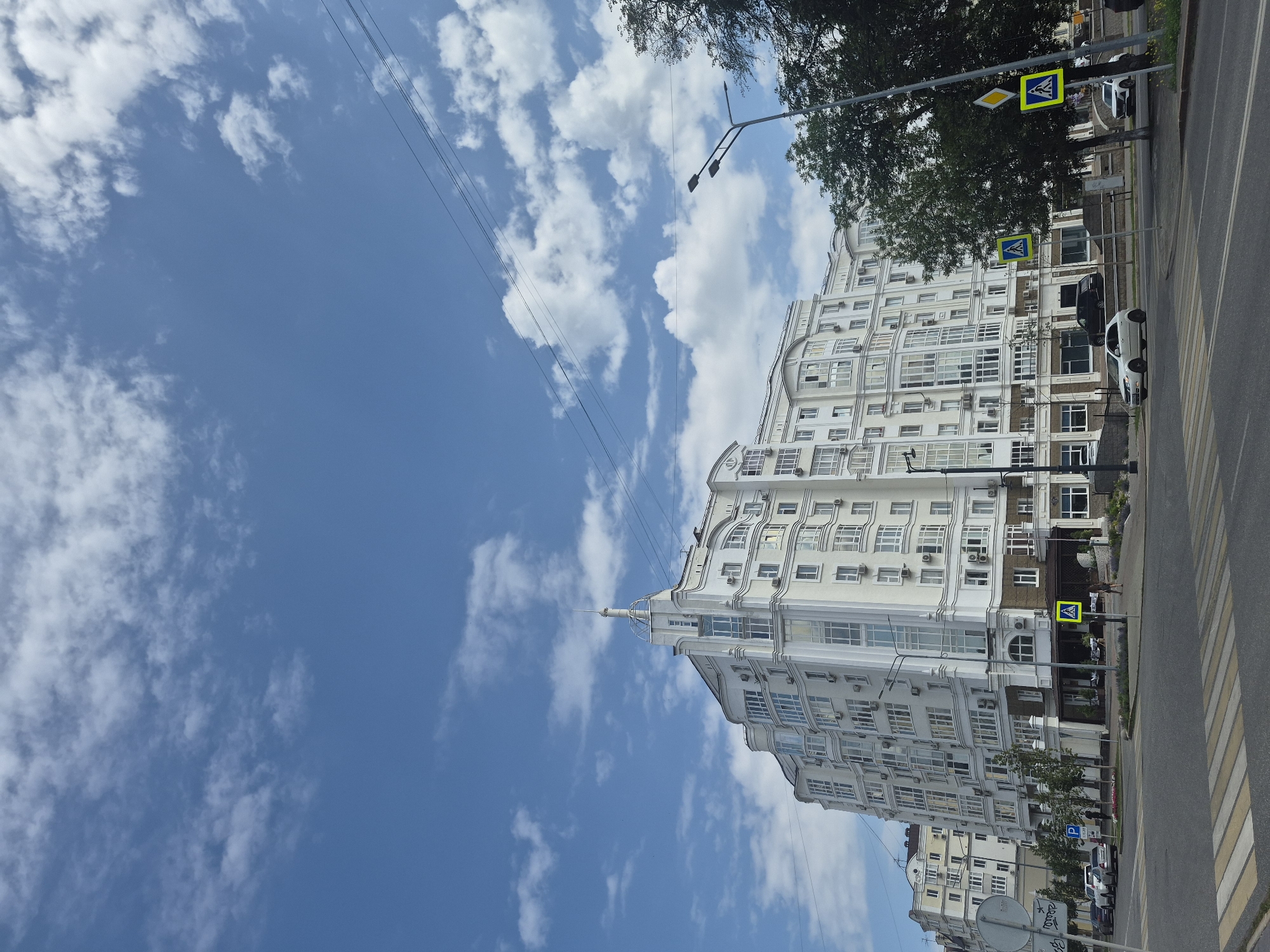 The image captures a bright, cloudy day in Ufa, Russia, with a large, modern, multi-story white building dominating the center. The building features numerous windows, balconies, and decorative architectural elements, suggesting it is an apartment complex or a commercial building. In the foreground, a street intersection is visible with a pedestrian crossing. Several cars are present, including a white sedan and a black SUV. People are seen walking on the sidewalk and near the intersection. Trees line the street, and streetlights are positioned along the roadside. The sky is a clear blue, interspersed with fluffy white clouds. The overall scene depicts an urban environment with contemporary architecture and typical city street activity.
