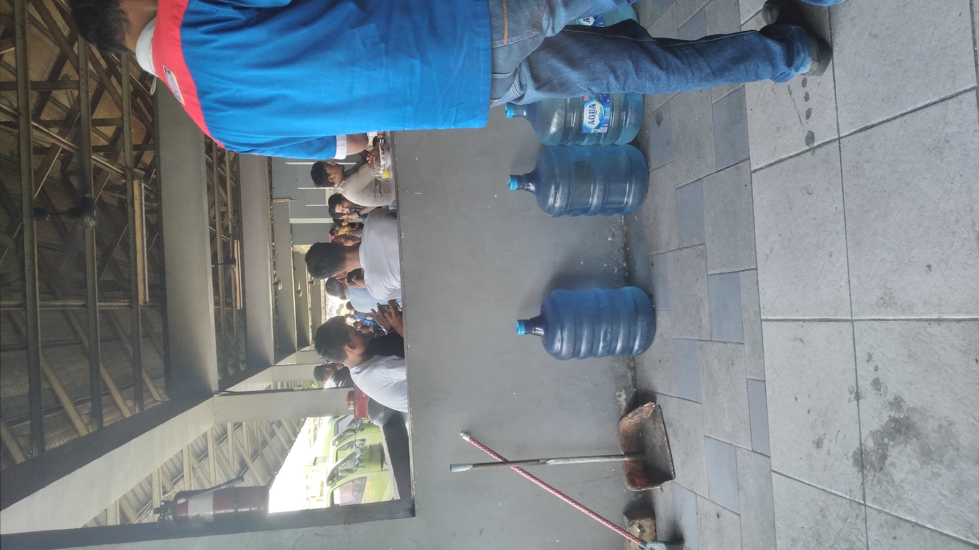 In a covered outdoor area in Purwakarta, Indonesia, a group of individuals is seated at tables, possibly dining. In the foreground, a person in a blue and red shirt and jeans stands facing away from the camera. To the left, a broom and dustpan rest against a gray wall. Lined up against the wall are three large blue water jugs. The area appears to be part of a business or service establishment, with a fire extinguisher visible on the left. In the background, several green vehicles are parked. The ceiling structure is visible above, supported by wooden beams.