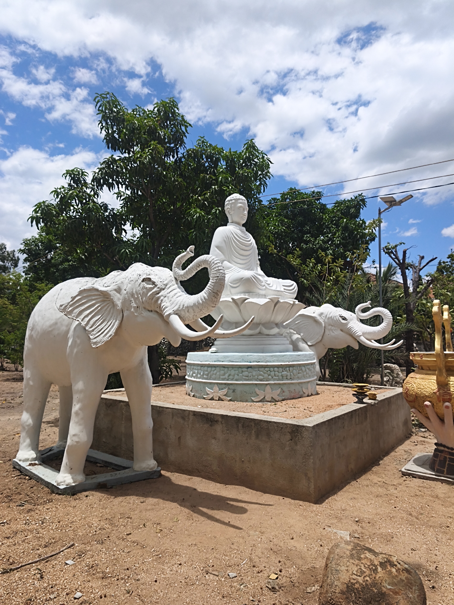 A statue of the Buddha sits on a lotus flower base, flanked by two white elephant statues, on a concrete pedestal in an outdoor, dirt and gravel area. The Buddha statue is white and shows the Buddha in a meditative pose. The elephant statues are also white and appear to be made of plaster or concrete. The one on the left is larger and in the foreground, facing left. The one on the right is smaller and partially obscured by the Buddha statue, facing right. Behind the statues is lush green foliage and trees under a blue sky with white clouds. A solar-powered street lamp is visible on the right side of the image. A golden vase or urn with decorative elements is also visible to the far right, held by a hand. The ground is covered with sandy soil and small stones, with some larger rocks scattered around.