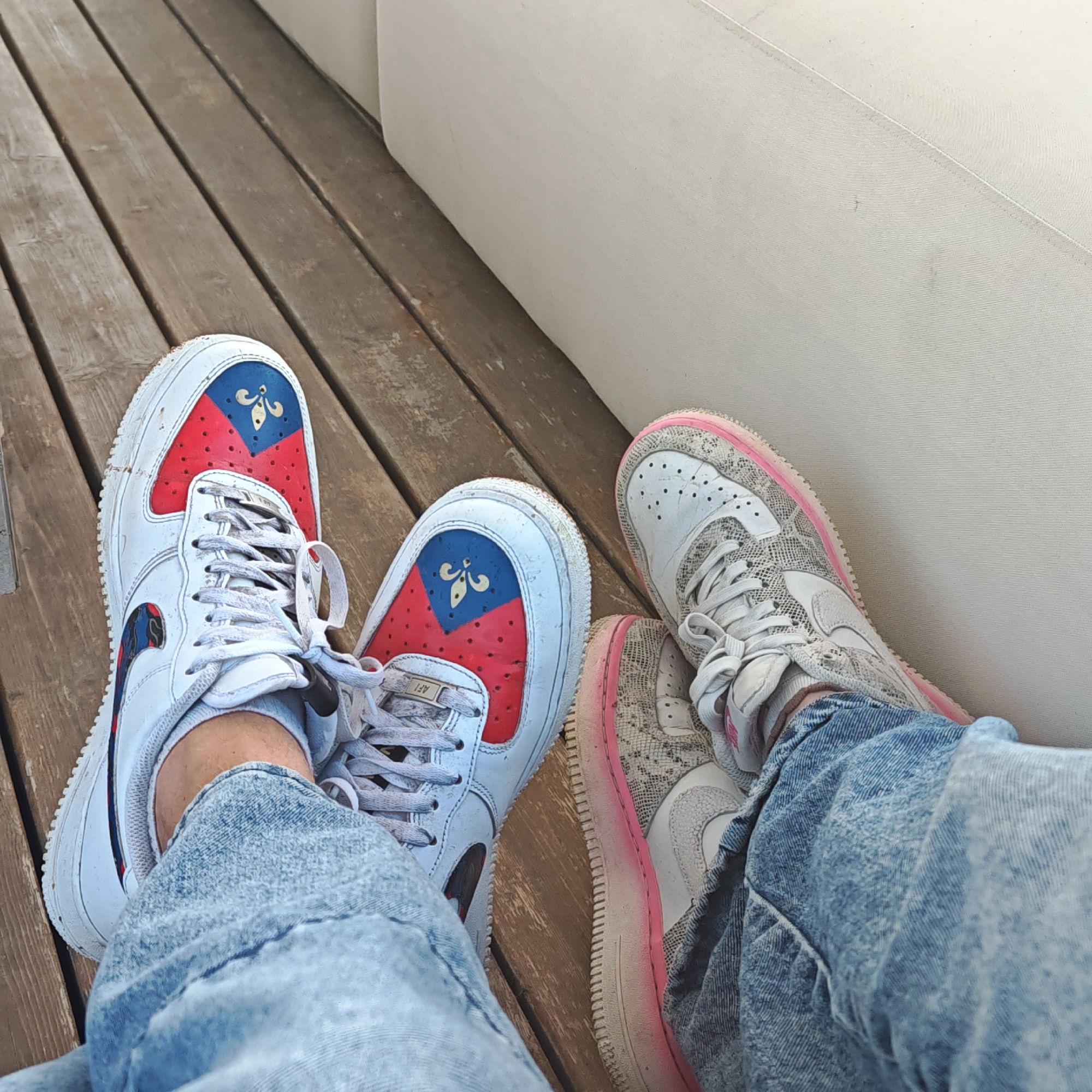 The image shows a pair of legs and feet resting on a wooden deck, likely a patio or similar outdoor setting. The left foot wears a white sneaker with a red and blue design on the toe, featuring a fleur-de-lis. The right foot is in a white and gray patterned sneaker with a pink-accented sole. Both individuals are wearing light-wash denim jeans. A light-colored, possibly white, upholstered surface or wall is visible in the background. The scene appears relaxed and casual. The location, Мысовское сельское поселение, Ukraine, cannot be visually confirmed but is information being requested.