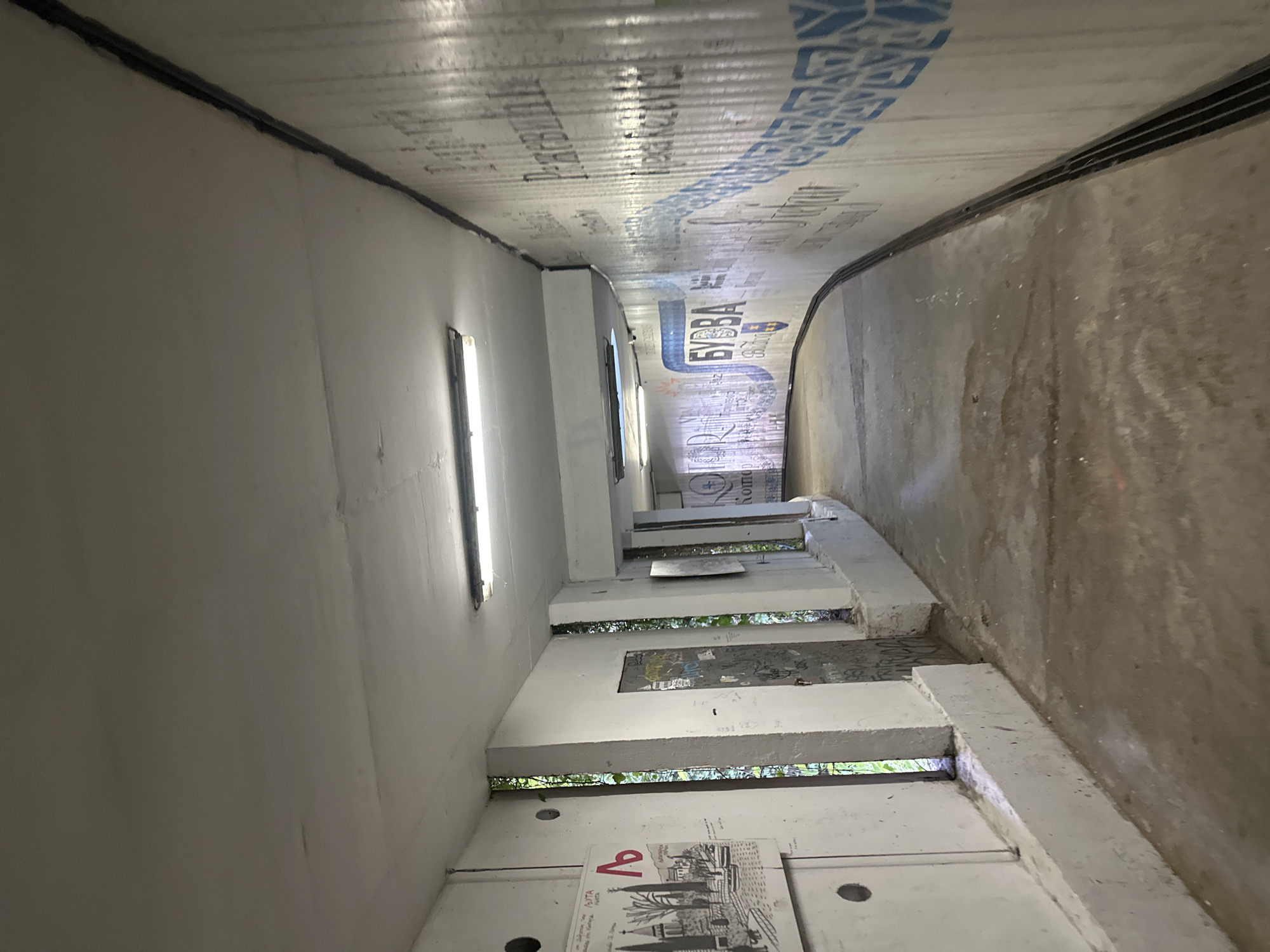 The image depicts a concrete passageway or underpass, captured with a tilted perspective that rotates the scene counter-clockwise. The left side of the frame shows a light-colored, possibly off-white, wall that curves gently into the distance. Along this wall, a series of concrete tiers or steps ascend towards the background. Gaps between these tiers reveal glimpses of green foliage and natural light, suggesting an opening to the outside. On the lowest visible tier, there is a piece of paper or a poster featuring a black-and-white sketch of buildings and trees, with a red "a" logo and Cyrillic text including the word "БУДВА" (Budva). Further along the tiers, another surface appears covered in graffiti. Fluorescent tube lights are mounted vertically on this wall, providing illumination.
The right side of the passageway features a textured, darker concrete wall, also curving into the distance. The ceiling of the underpass is covered with a reflective material, possibly corrugated plastic or metal, adorned with blue patterns and Cyrillic text. Visible words on the ceiling include "БИВБА" (BIVBA) and "KONTO", along with other partial or reversed characters.
The passage recedes into the background, creating a tunnel-like effect. The combination of artificial lighting and natural light filtering through the openings suggests the photograph was taken during the day. No people are visible. The scene portrays an urban environment, specifically located in Budva, Montenegro, consistent with the visible text.
