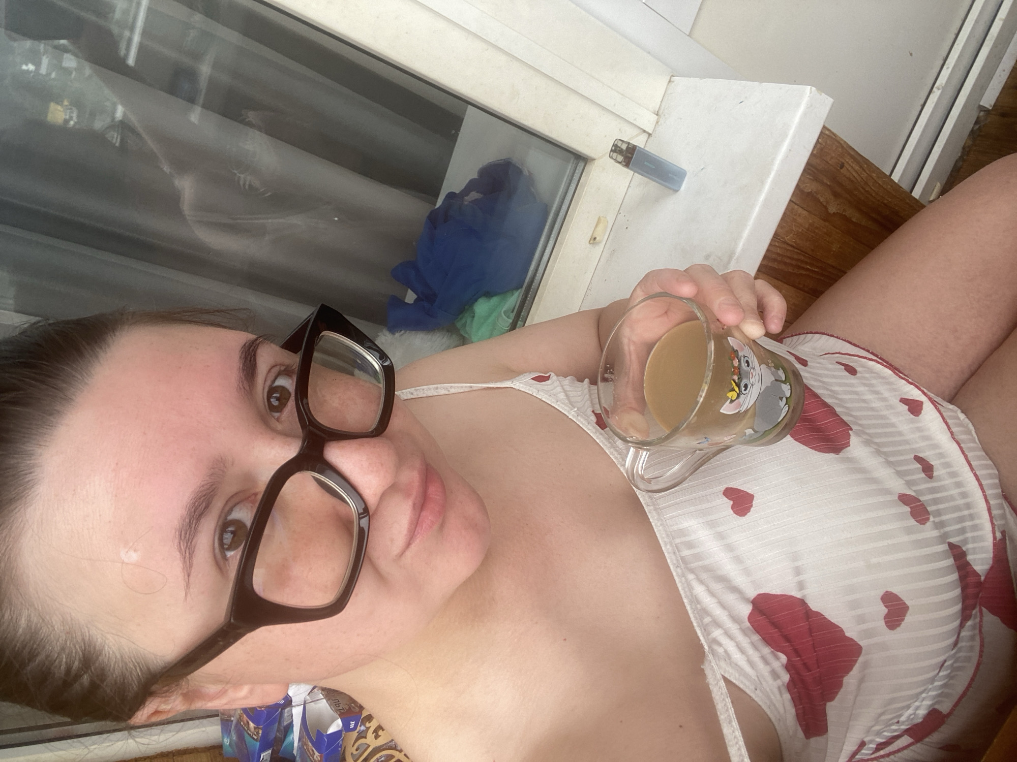 A woman wearing glasses and a white camisole with red hearts holds a mug filled with a light brown beverage. She is seated indoors, near a window. A blue vape pen rests on the windowsill next to her. The background shows clothing items draped over a ledge and a reflection in the glass. The image is a close-up shot of the woman's upper body and the items she is holding. There is no indication of the city of Chaikovsky, Russia, or any specific urban environment.