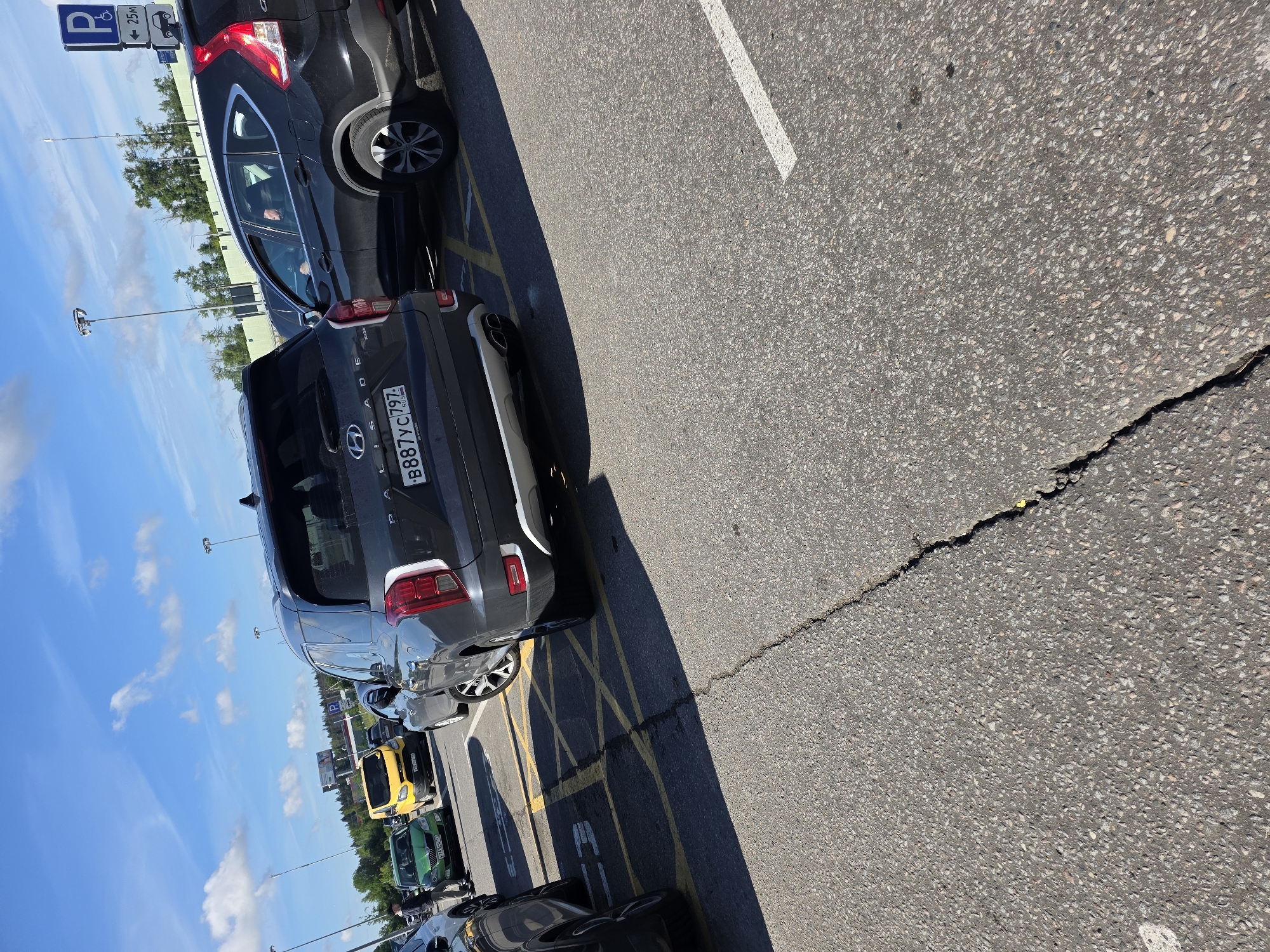 In an outdoor, paved parking lot under a bright, partly cloudy sky, several vehicles are parked. The primary focus is a dark gray Hyundai Palisade SUV, seen from the rear, with its license plate reading "B887YC 797 RUS". To its right, a black Honda CR-V SUV is partially visible, with a person in the driver's seat. Further left, a green hatchback is parked, and a bright yellow van is behind it. The parking lot surface is asphalt with visible cracks and yellow painted markings, including a diagonal crosshatch pattern indicating a no-parking zone. In the background, trees line the horizon, and utility poles with lights are scattered throughout the area. A blue and white parking sign with an accessibility symbol and distance marker is visible on a pole in the upper right quadrant.
