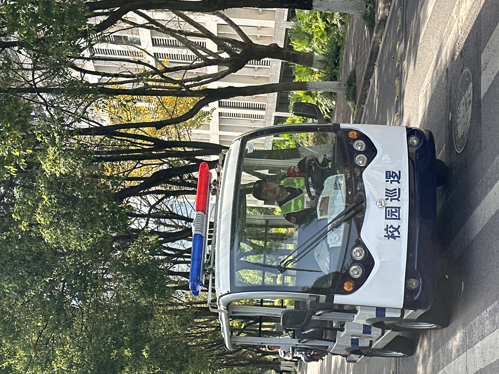 A white and blue electric campus patrol vehicle is depicted driving on a tree-lined road under bright daylight. The vehicle features a white front merging into a dark blue body, with red and blue emergency lights mounted on its roof. Inside, a person wearing a green high-visibility vest is visible in the driver's seat, holding the steering wheel. Another individual is partially seen in the rear of the vehicle.
The road surface includes speed bumps and a circular manhole cover. Lush green trees with some yellowing foliage line both sides of the road, suggesting a verdant environment, possibly during autumn. Behind the trees, modern multi-story buildings with balconies are visible, indicating an institutional setting like a university campus in Jiangjin, China. The bright sunshine suggests clear weather and daytime.
Prominently displayed on the side of the vehicle in vertical Chinese characters is "校园巡逻" (Xiàoyuán Xúnluó), which translates to "Campus Patrol," clearly indicating its function within the campus environment. The overall scene conveys a sense of routine security or maintenance activity on a well-kept campus.