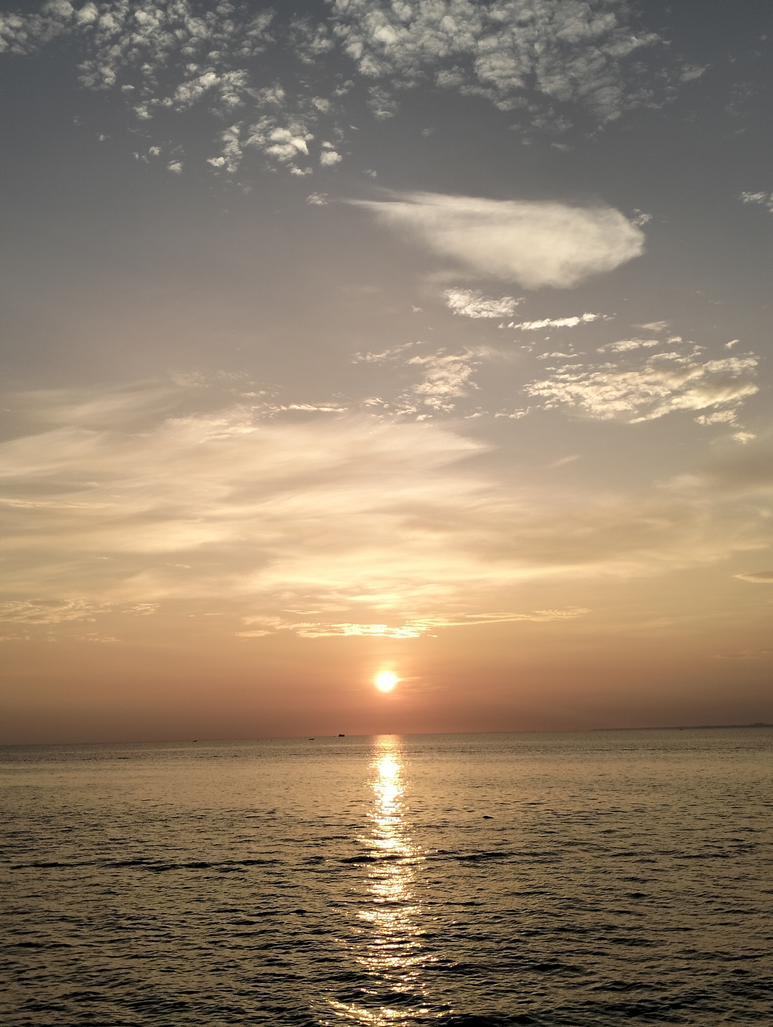 The image depicts a sunset over the ocean, likely in Lhokseumawe, Indonesia, given the prompt. The sun is positioned low on the horizon, casting a golden glow across the water and the lower portion of the sky. The reflection of the sun creates a bright, shimmering path on the water's surface. The sky above is a gradient of colors, ranging from a light, warm tone near the horizon to a cooler, grayish tone higher up. Clouds are scattered throughout the sky, with some dense patches at the top and more diffuse formations lower down. The water is relatively calm, with gentle ripples visible across its surface. There are no people visible in the image, but small vessels are present in the far distance. The overall scene conveys a sense of tranquility and natural beauty.