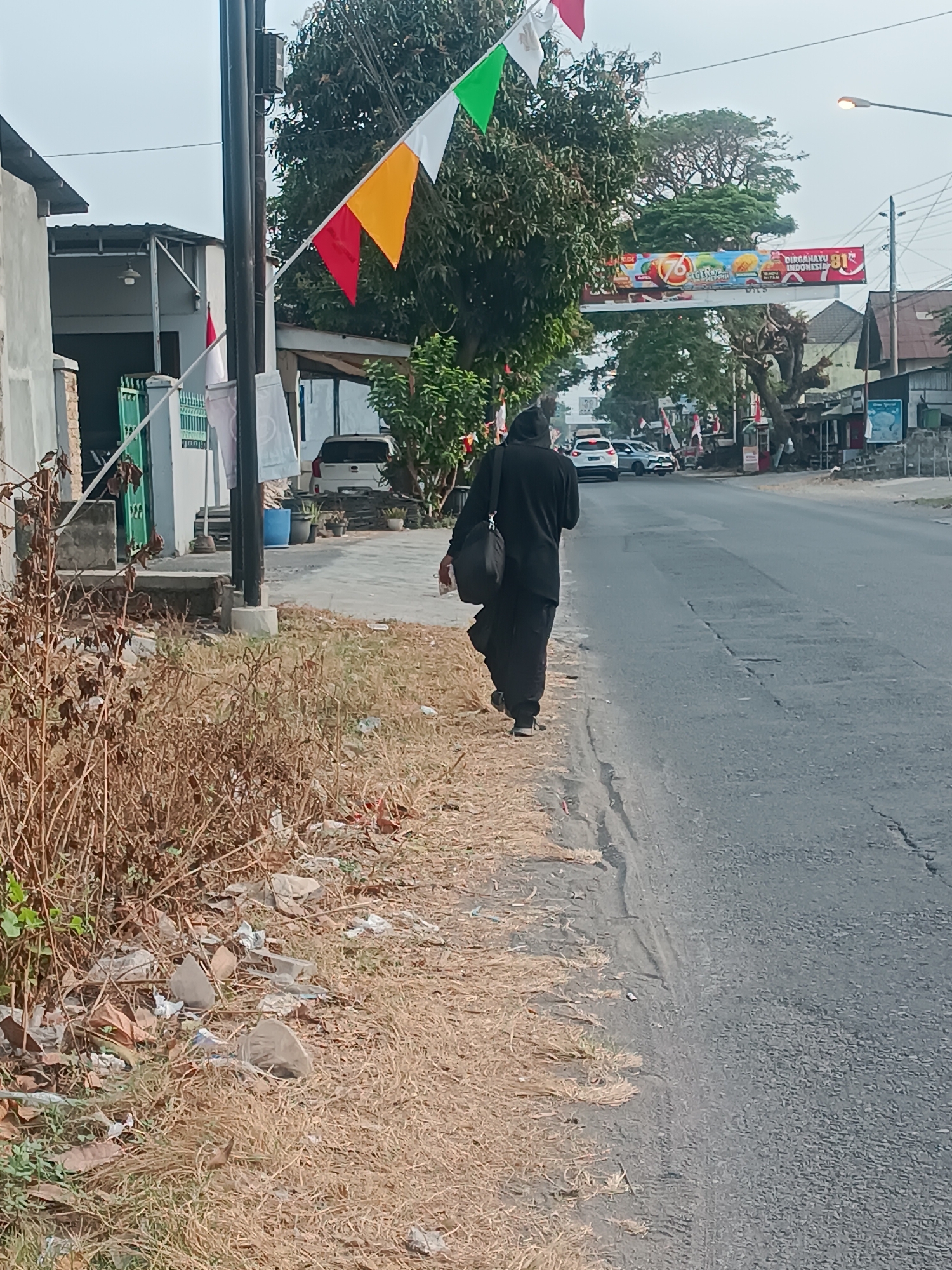 A person wearing a black hijab and robe walks on the right side of a paved road in Sewon, Indonesia. The person carries a black bag over their left shoulder. The road has some cracks and debris. To the left of the road is a dry, grassy area with scattered litter and some dead bushes. A white and grey building is visible on the far left, with a green gate. In the background, across the street, are more buildings, trees, and a banner with Indonesian text and graphics. There are several cars on the road and a street lamp is visible. Small triangular flags in red, yellow, and green are strung across poles in the foreground. The sky is overcast.